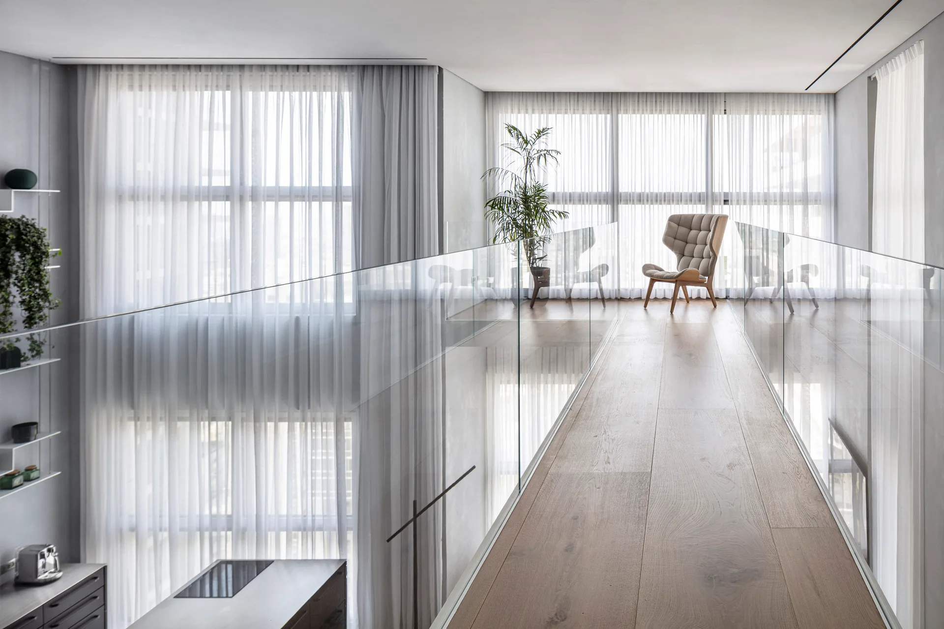 A minimalist mezzanine walkway features light wood plank flooring, frameless glass railings, and a grey tufted armchair by sheer-curtained windows.