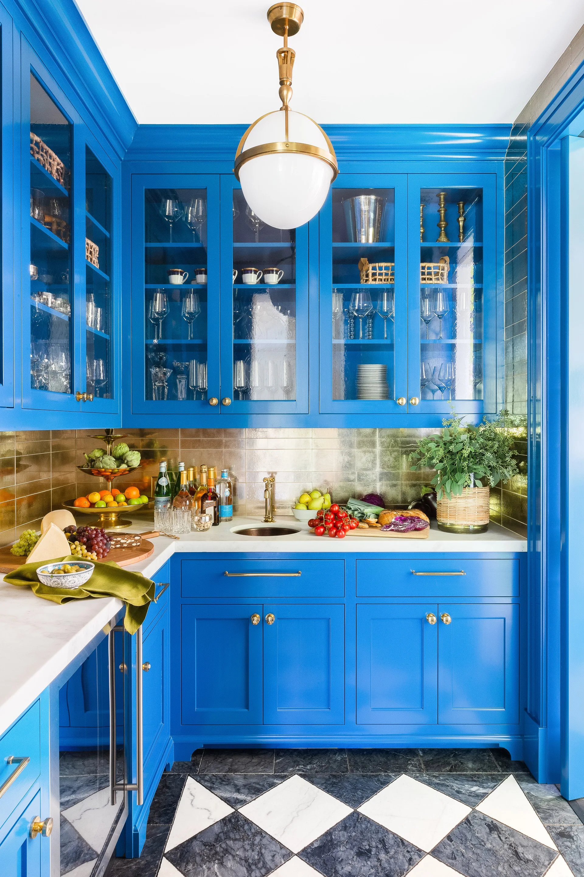 Bright blue kitchen cabinets feature glass door fronts, a gold metallic tile backsplash, white countertops, and a black and white checkerboard floor.