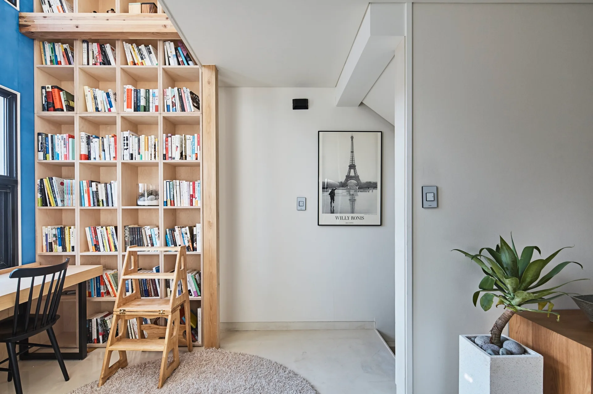 A home library features a floor-to-ceiling light wood bookshelf filled with books, a matching wooden desk with a black chair, and a wooden stepladder.