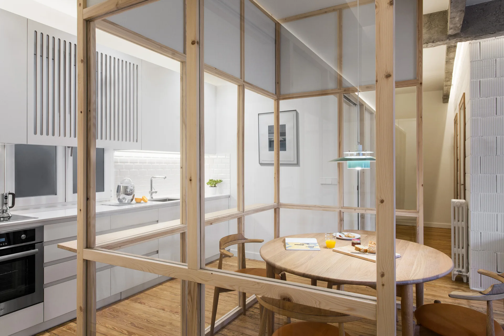 A modern kitchen with white cabinets is separated from a round wood dining table by a light wood framed glass partition.