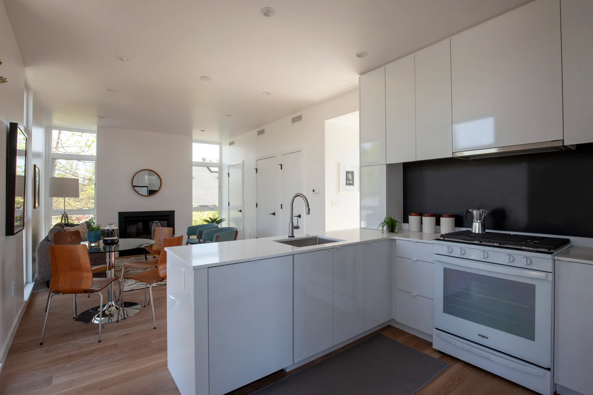A modern open-plan kitchen with high-gloss white cabinets, a white island, a black backsplash, and an adjacent dining area.