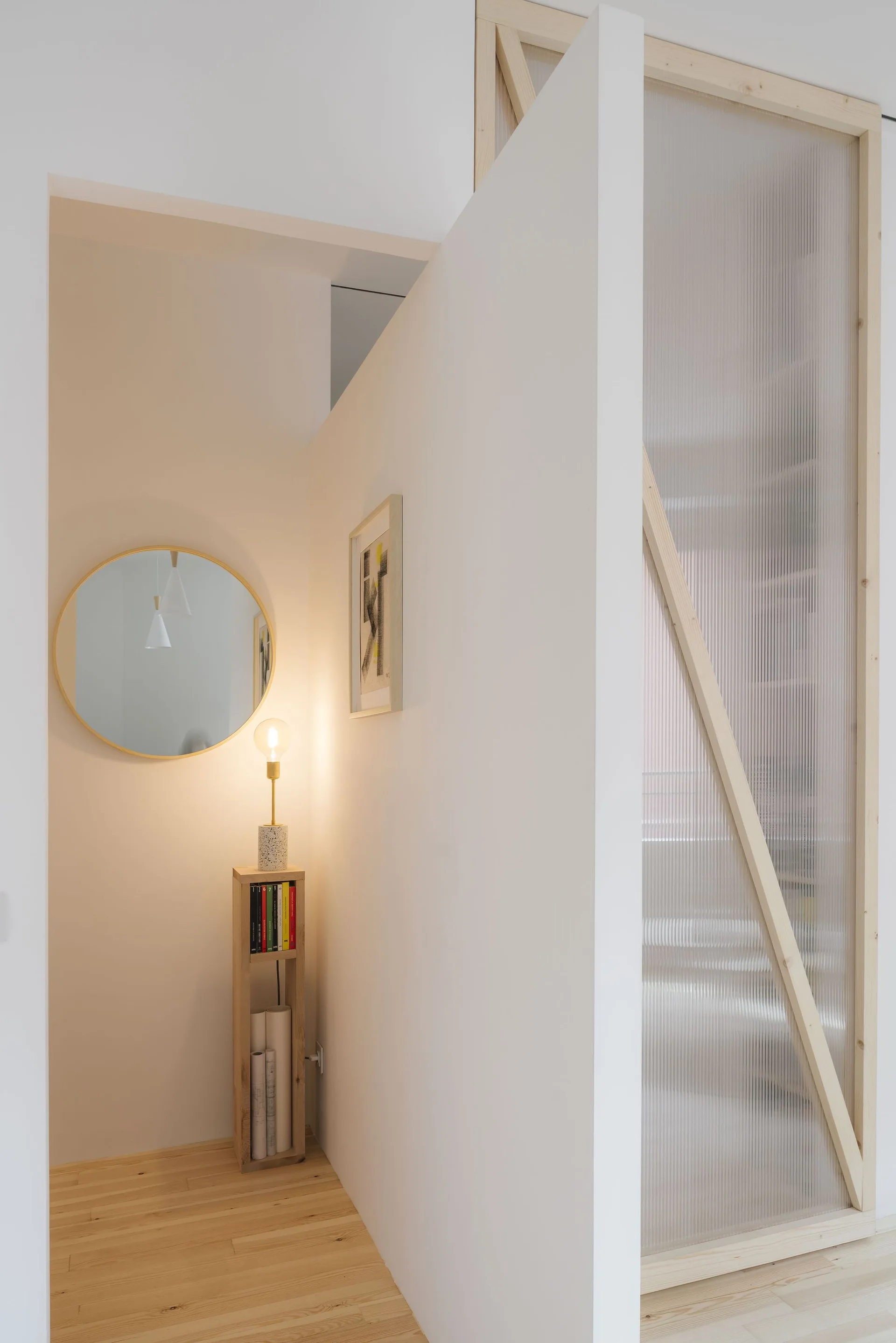 A minimalist hallway with a round mirror, a slender wooden shelf holding a lamp, and a tall fluted glass room partition.