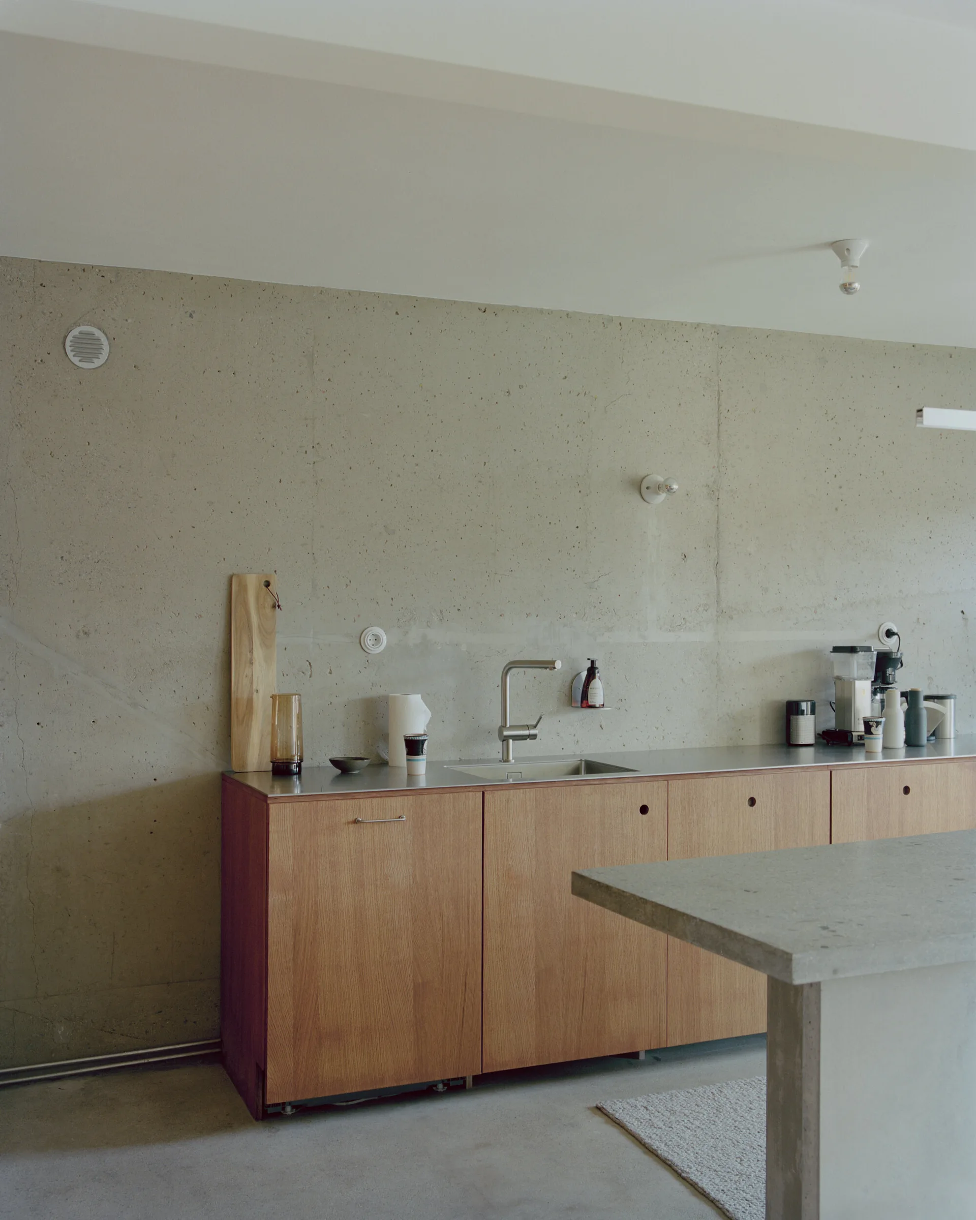 A minimalist kitchen features light wood cabinets, a metal countertop, and an undermount sink against a raw grey concrete wall.