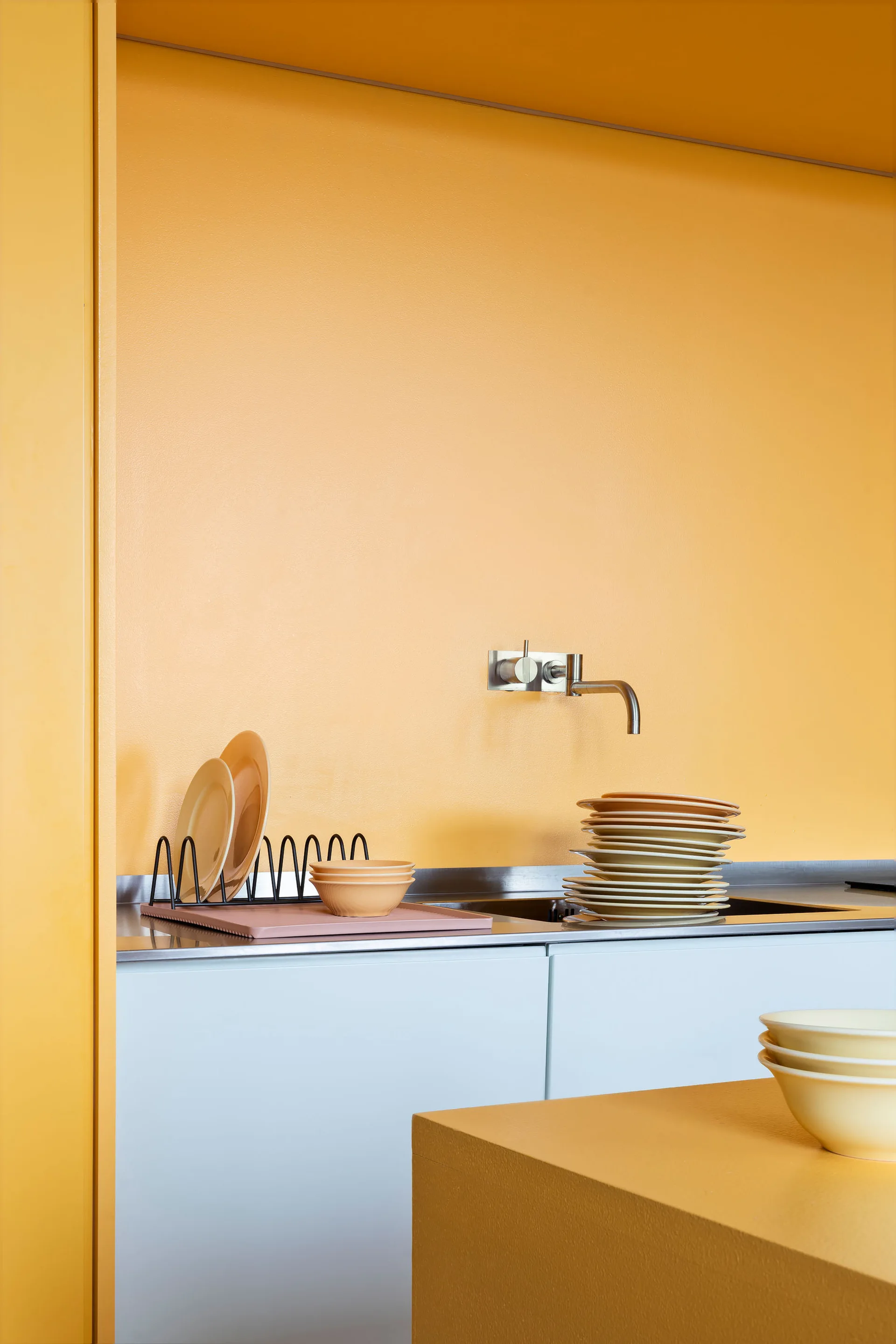 A minimalist kitchen features a bright yellow wall, light grey lower cabinets, a wall-mounted steel faucet, and stacks of plates by the sink.