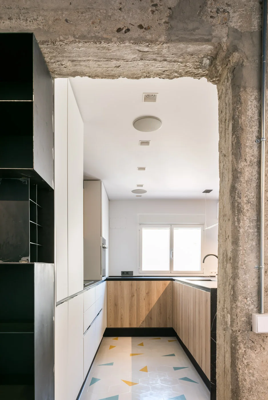 A modern kitchen with wood and white cabinetry and patterned hexagon floor tiles, viewed through a rough concrete structural opening.