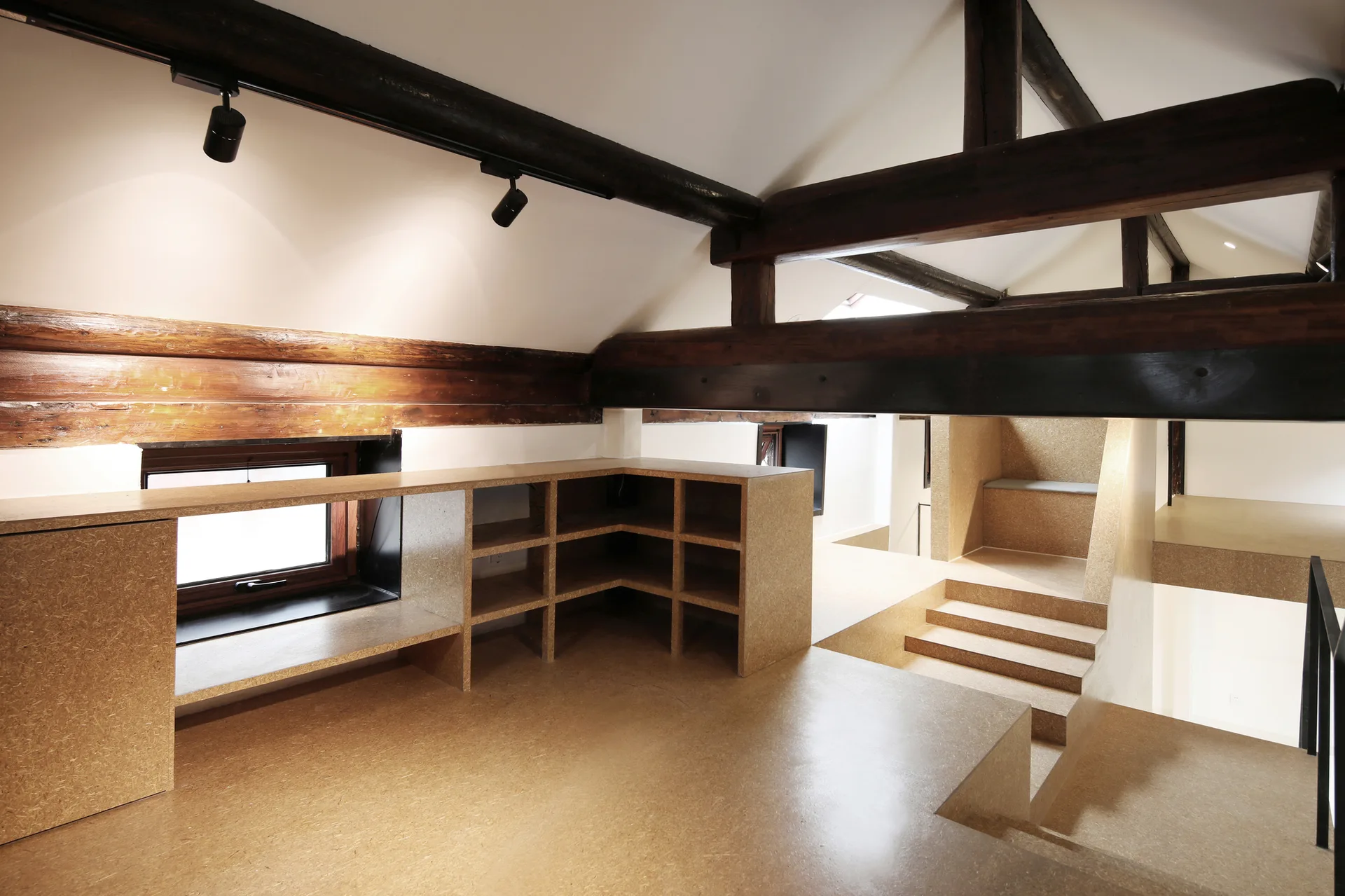 A multi-level attic loft features exposed dark timber beams, a white vaulted ceiling, and built-in shelving constructed from oriented strand board.