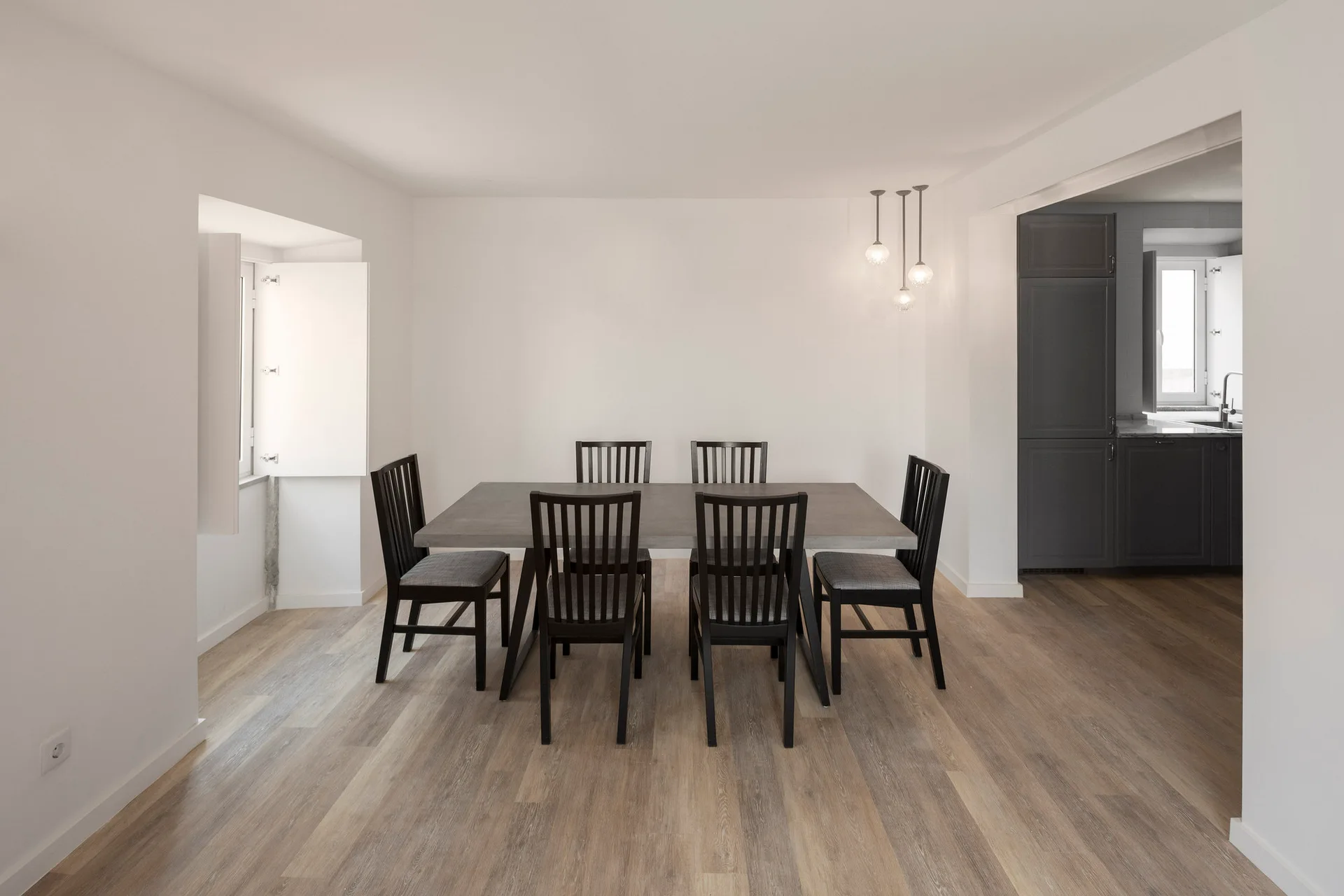 A minimalist white dining room features a gray rectangular table and six black spindle-back chairs on light wood flooring.