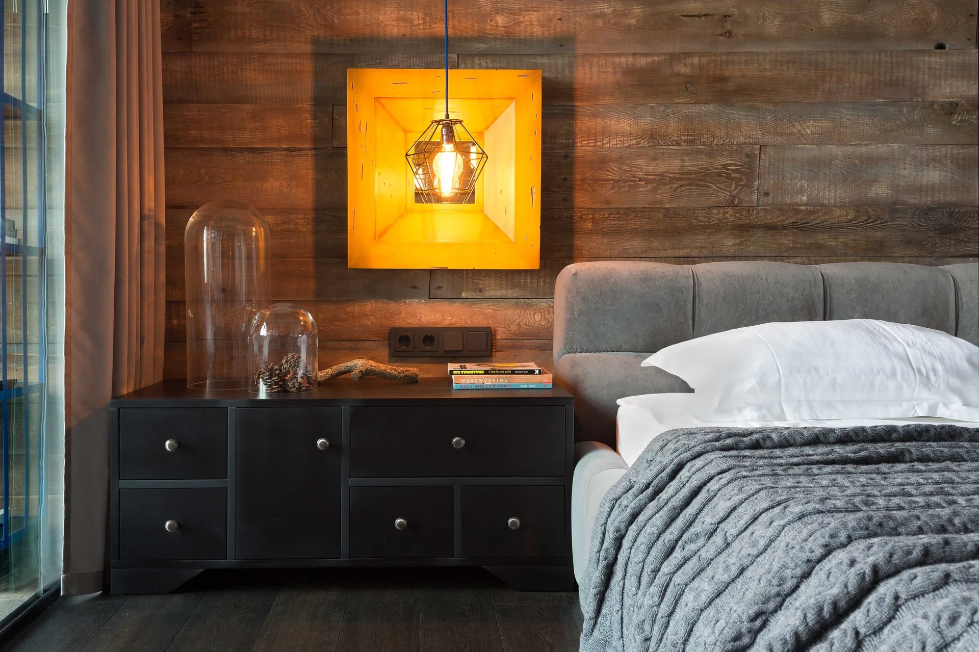 A rustic bedroom features a dark wood-paneled wall, a black nightstand with glass cloches, and a grey upholstered bed under a yellow pendant light.