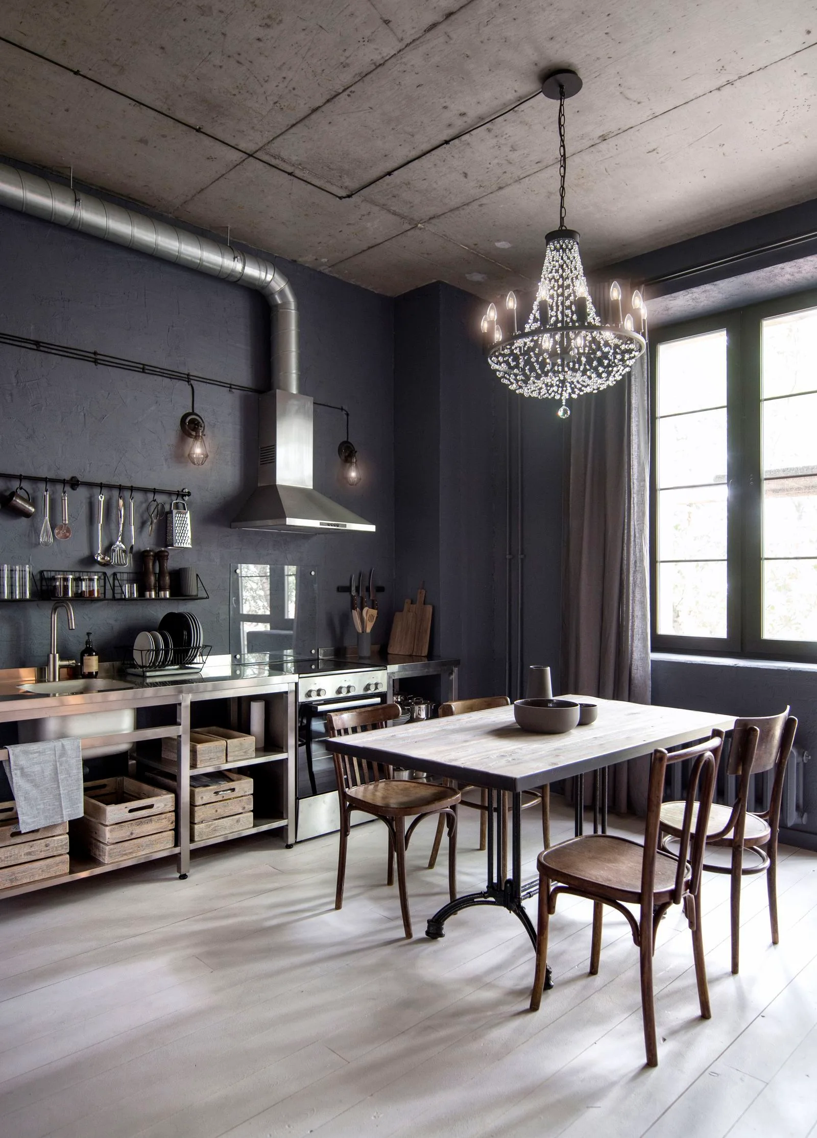 An industrial kitchen with dark charcoal walls, an exposed concrete ceiling, a stainless steel workstation, and a dining table under a crystal chandelier.