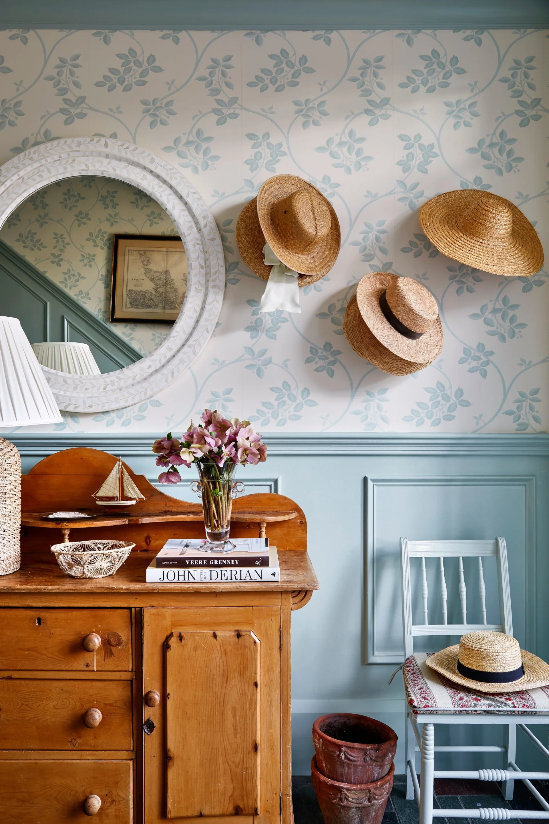 A wooden sideboard sits against light blue wainscoting and floral wallpaper decorated with hung straw hats, a round mirror, and a blue painted chair.