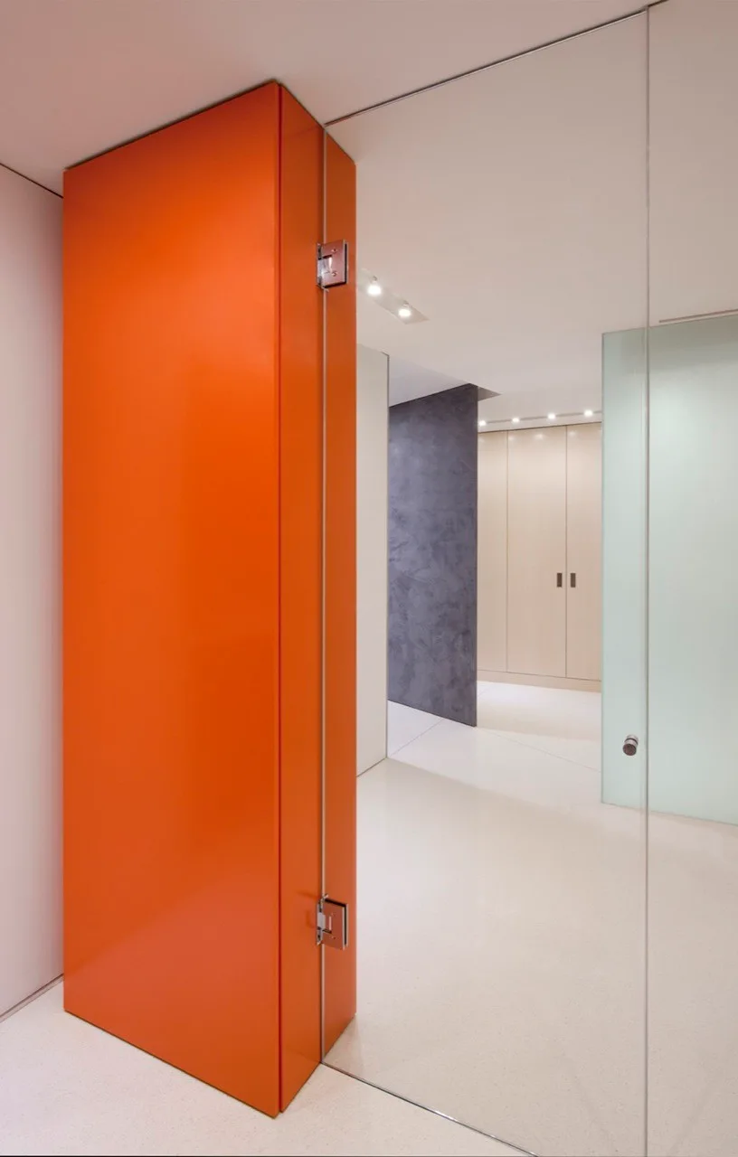A minimalist hallway features a glossy bright orange accent door alongside a glass partition, white flooring, and light wood built-in cabinets.