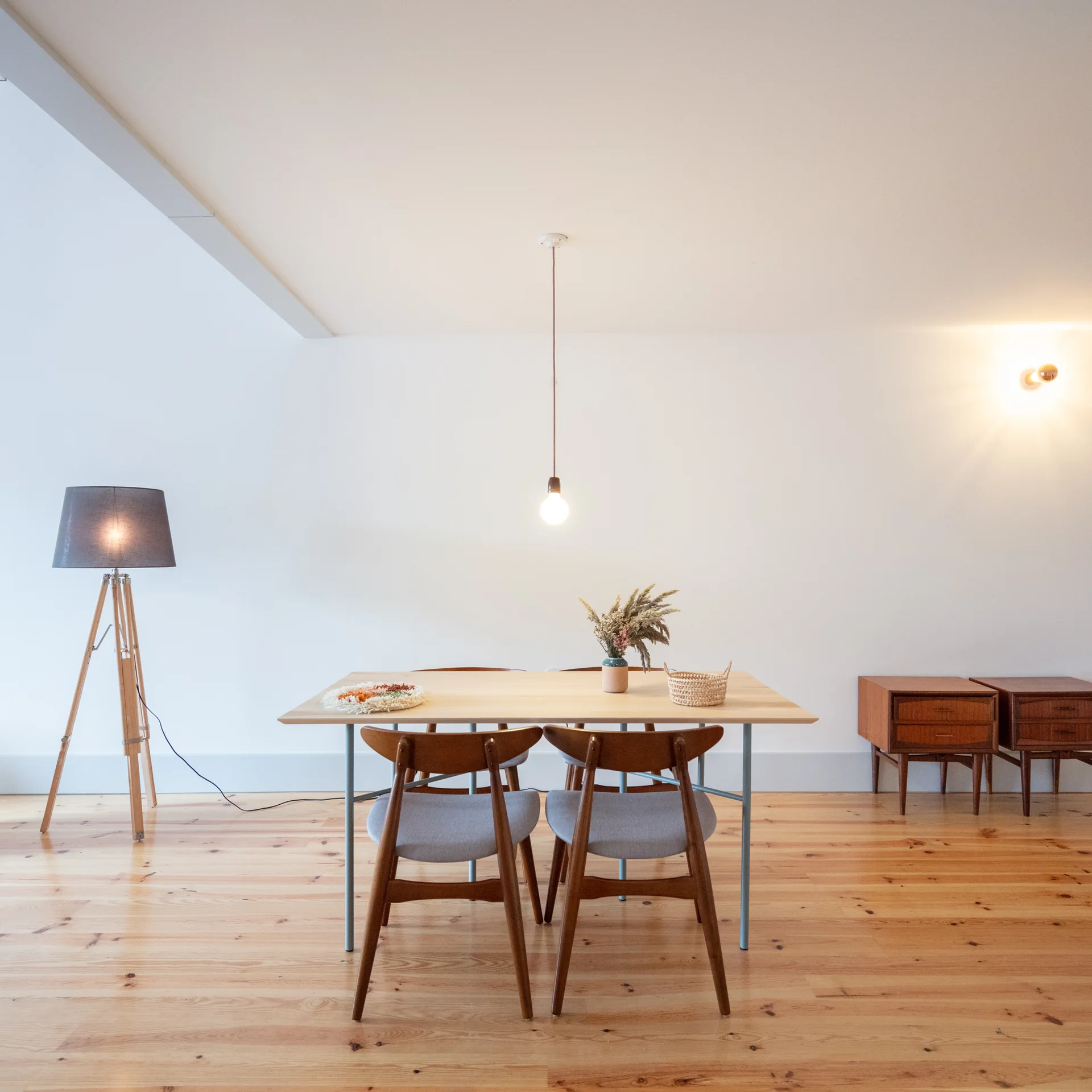 A minimalist dining room features a light wood table, mid-century wooden chairs, a tripod floor lamp, and warm hardwood flooring against plain white walls.