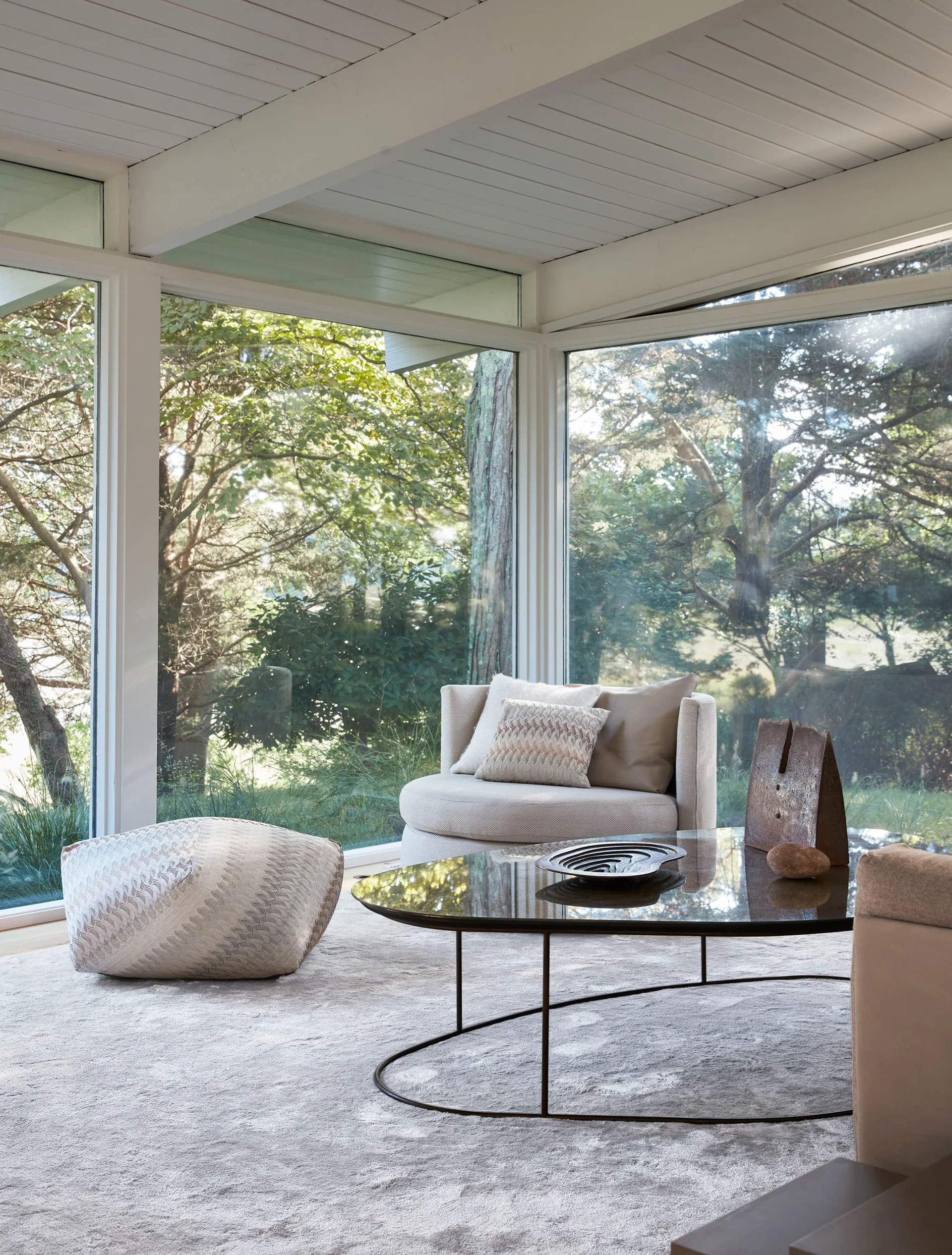 A sunlit living area features a round upholstered armchair, a low glass coffee table, and a textured pouffe set against large windows overlooking trees.