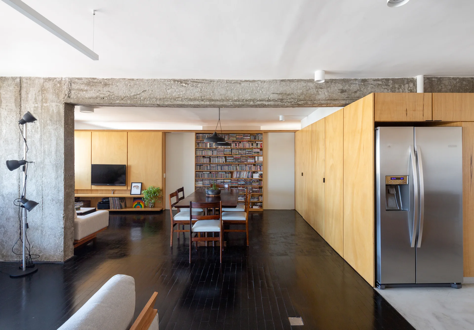 An open living and dining area featuring an exposed concrete beam, dark wood flooring, and extensive light wood built-in cabinetry.