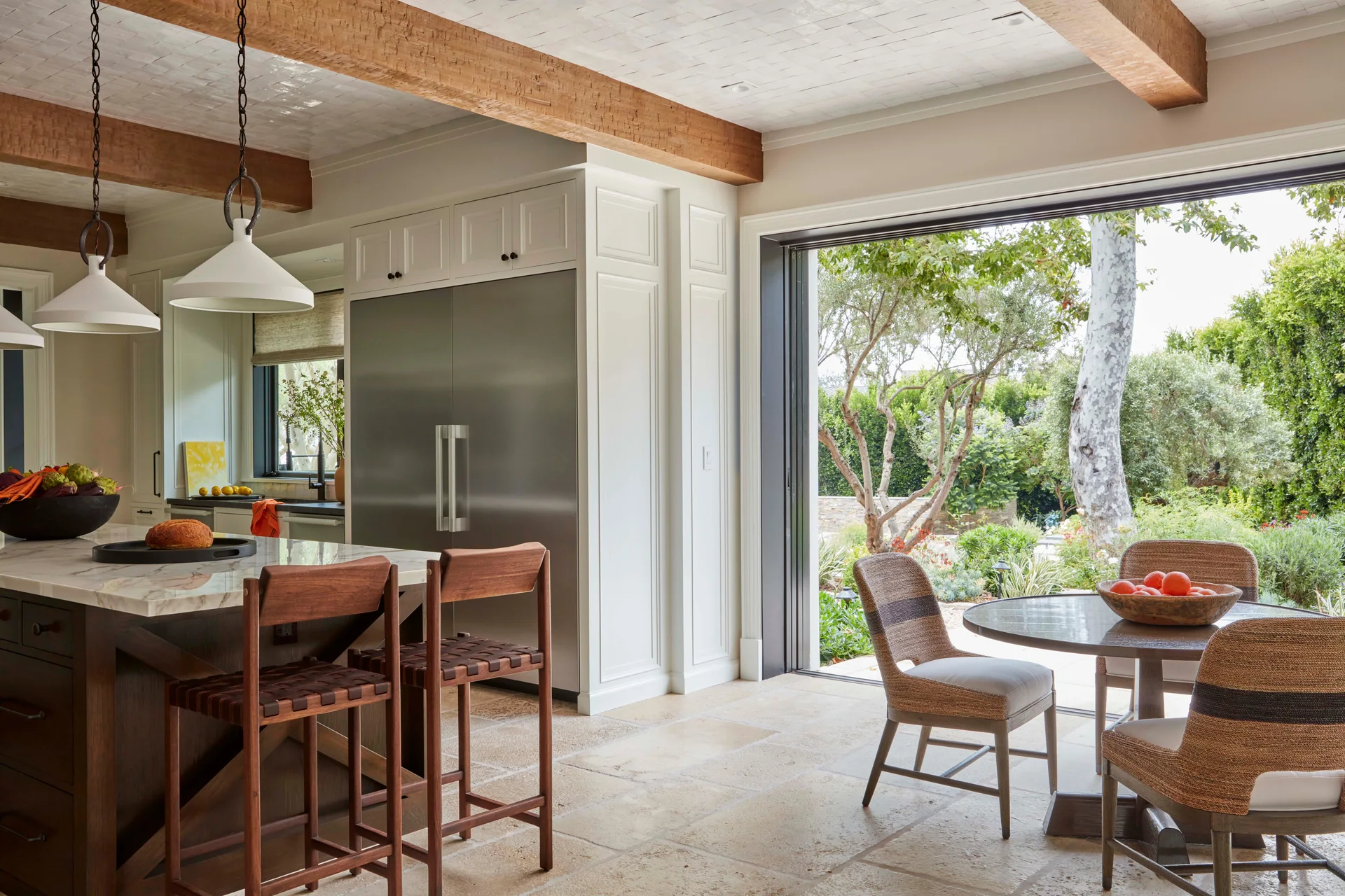 A bright kitchen with a marble island, wooden ceiling beams, stone tile floors, and a wide open folding door leading to a garden.