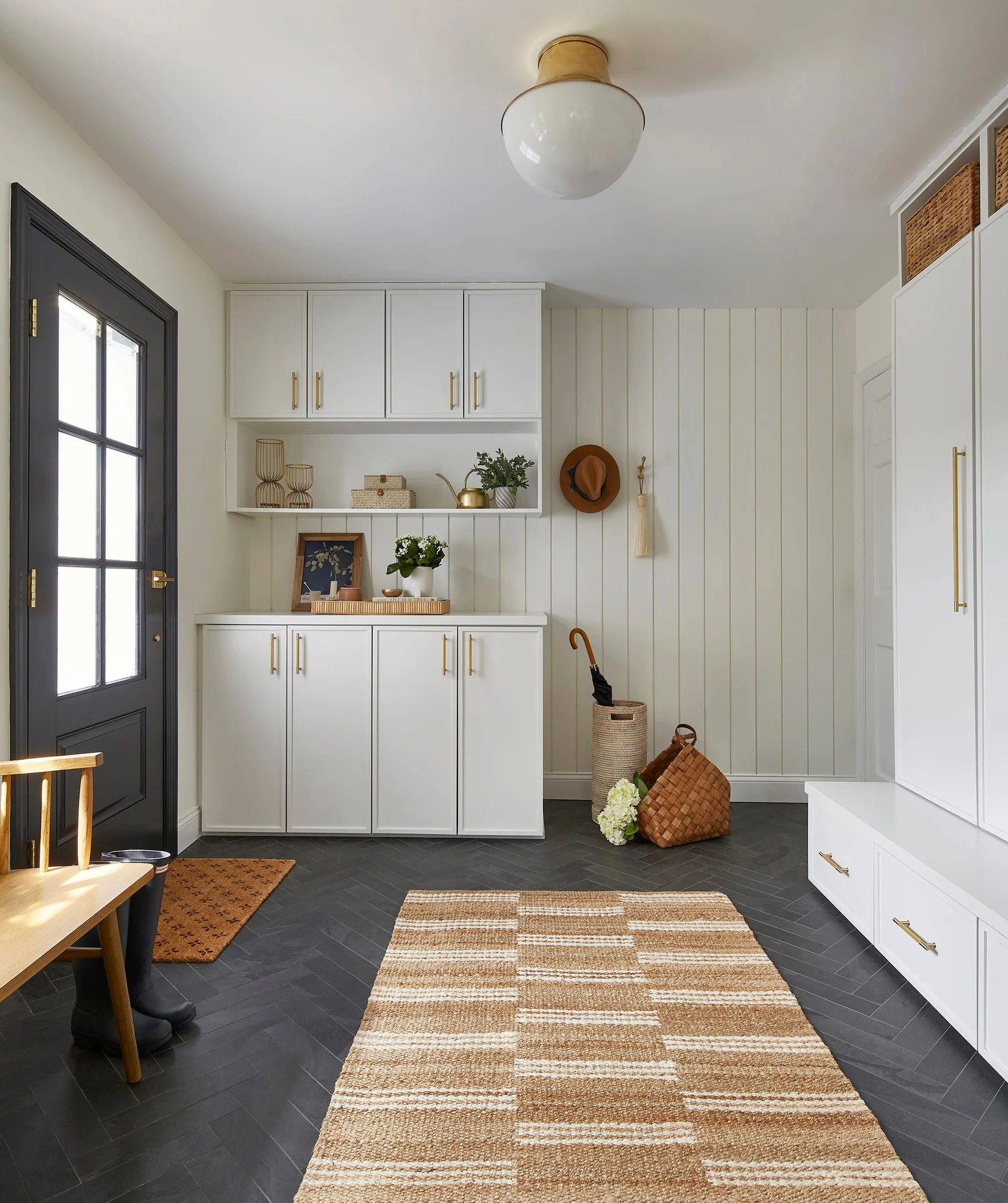 A bright mudroom with white built-in cabinets, a dark grey entry door, vertical wood wall paneling, and dark herringbone tile flooring.