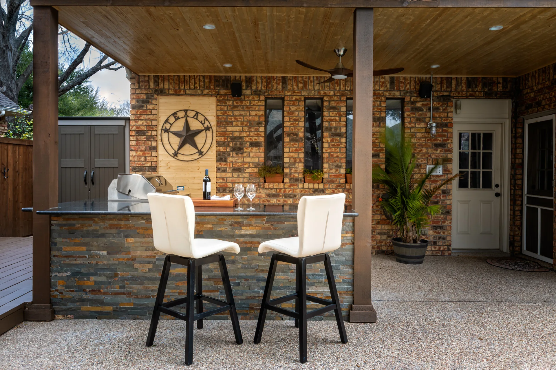A covered outdoor kitchen features a stone-faced bar with two white bar stools under a warm wood-paneled ceiling.