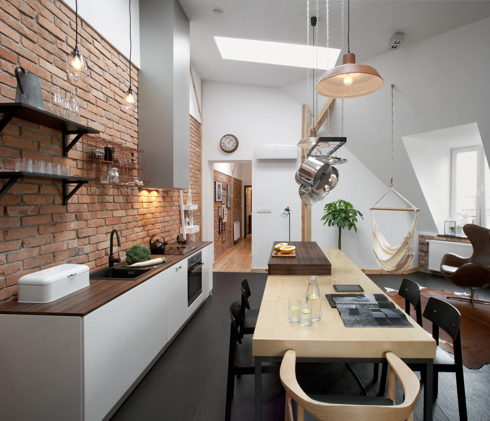 An industrial loft kitchen featuring a long exposed brick wall, white base cabinets, a light wood dining table with black chairs, and a skylight.