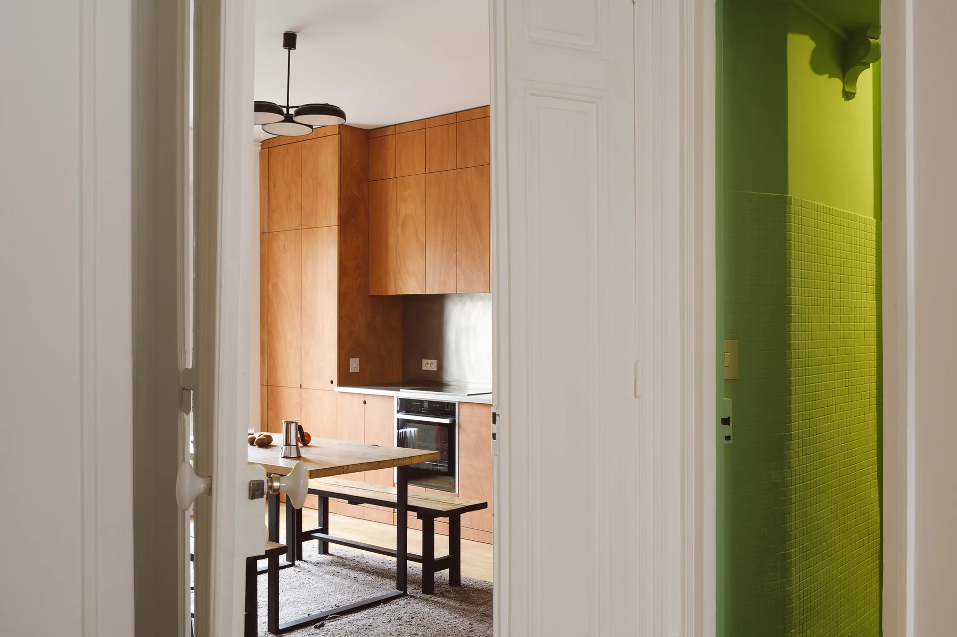 A view through a doorway showing warm wood plywood kitchen cabinets and a dining table next to a small area with bright green tiled walls.