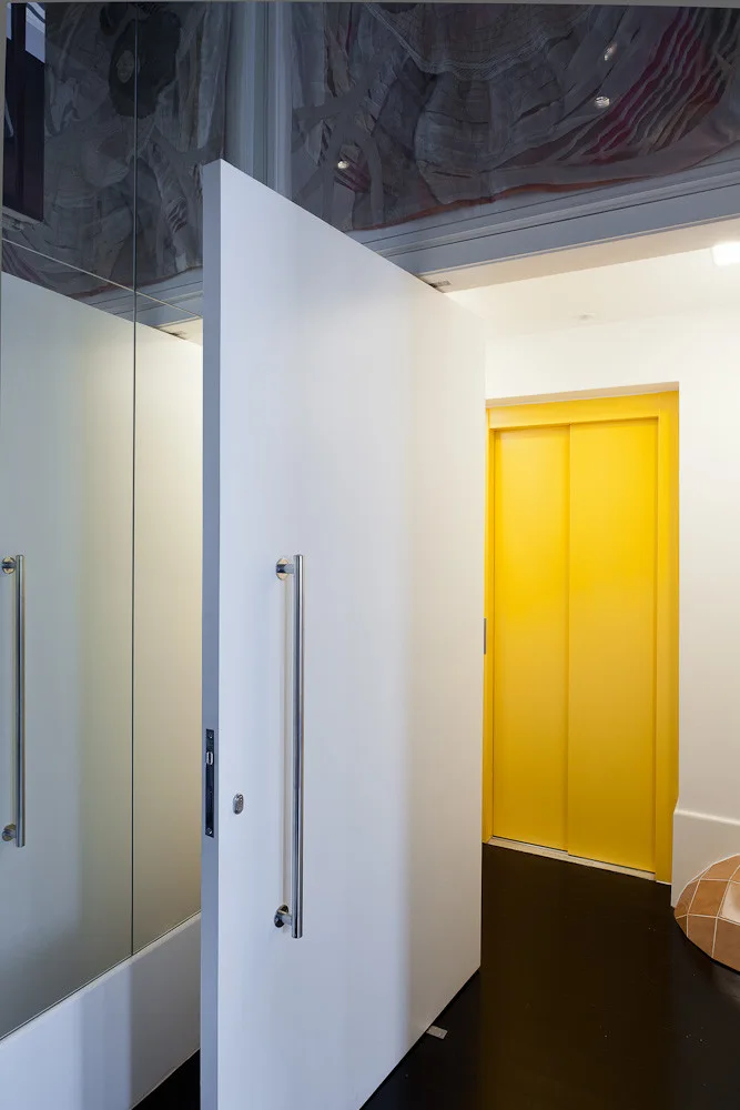 A modern entryway features a large white pivoting door with a long metal handle, a mirrored wall, and a bright yellow sliding door in the background.