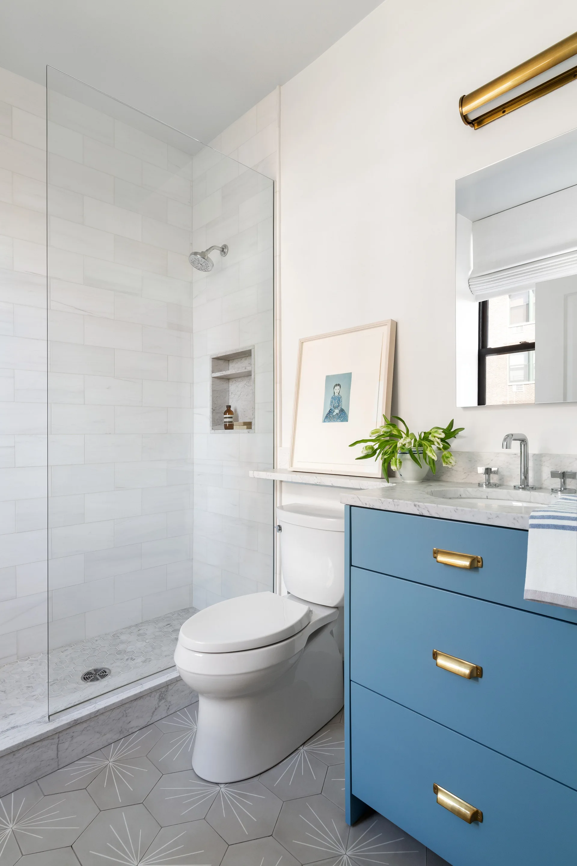 A modern bathroom features a slate blue vanity with brass drawer pulls, a white toilet, and a glass-enclosed shower with grey starburst floor tiles.