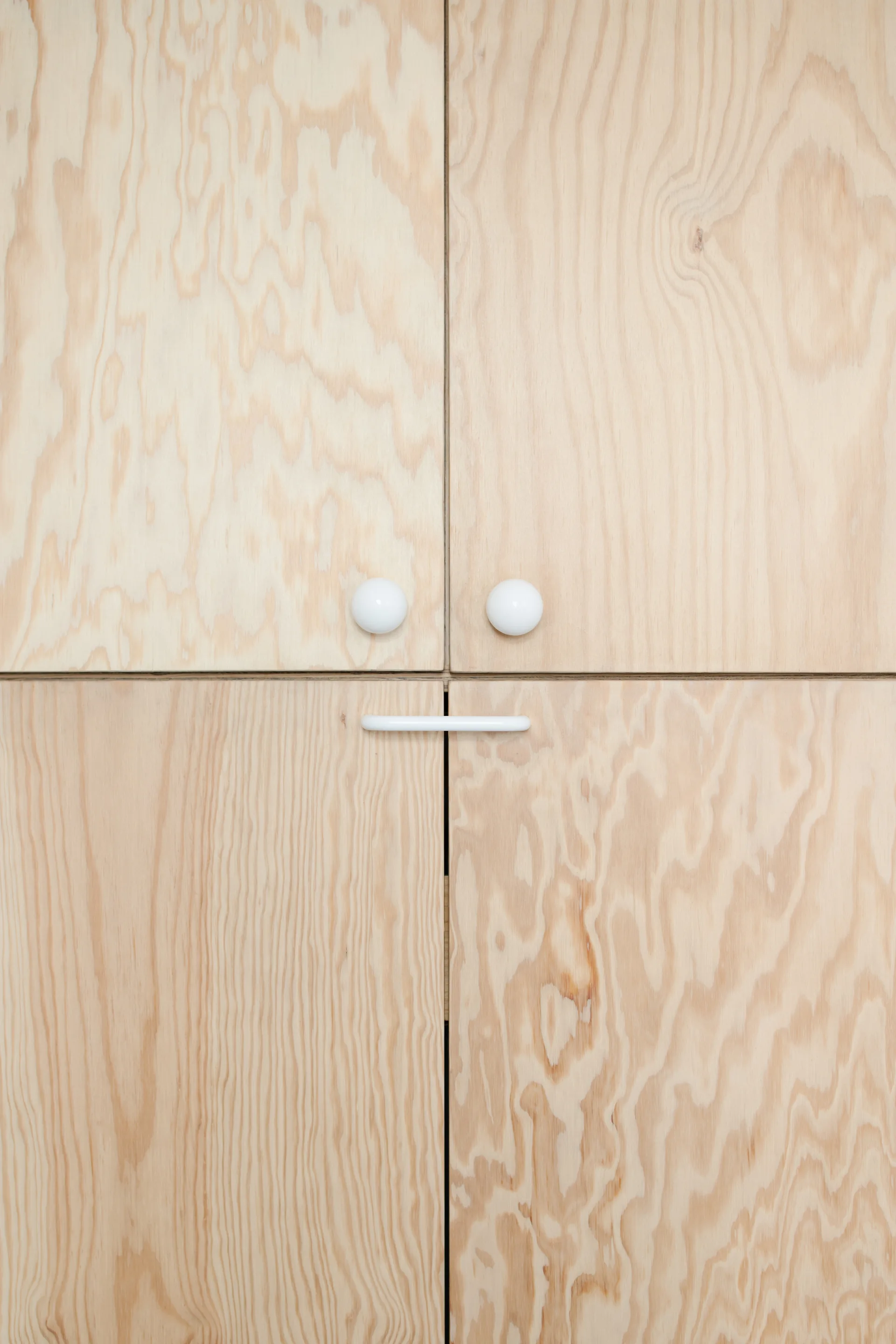 A close-up of light plywood cabinet doors featuring natural wood grain patterns and minimalist white knobs and a pull handle.