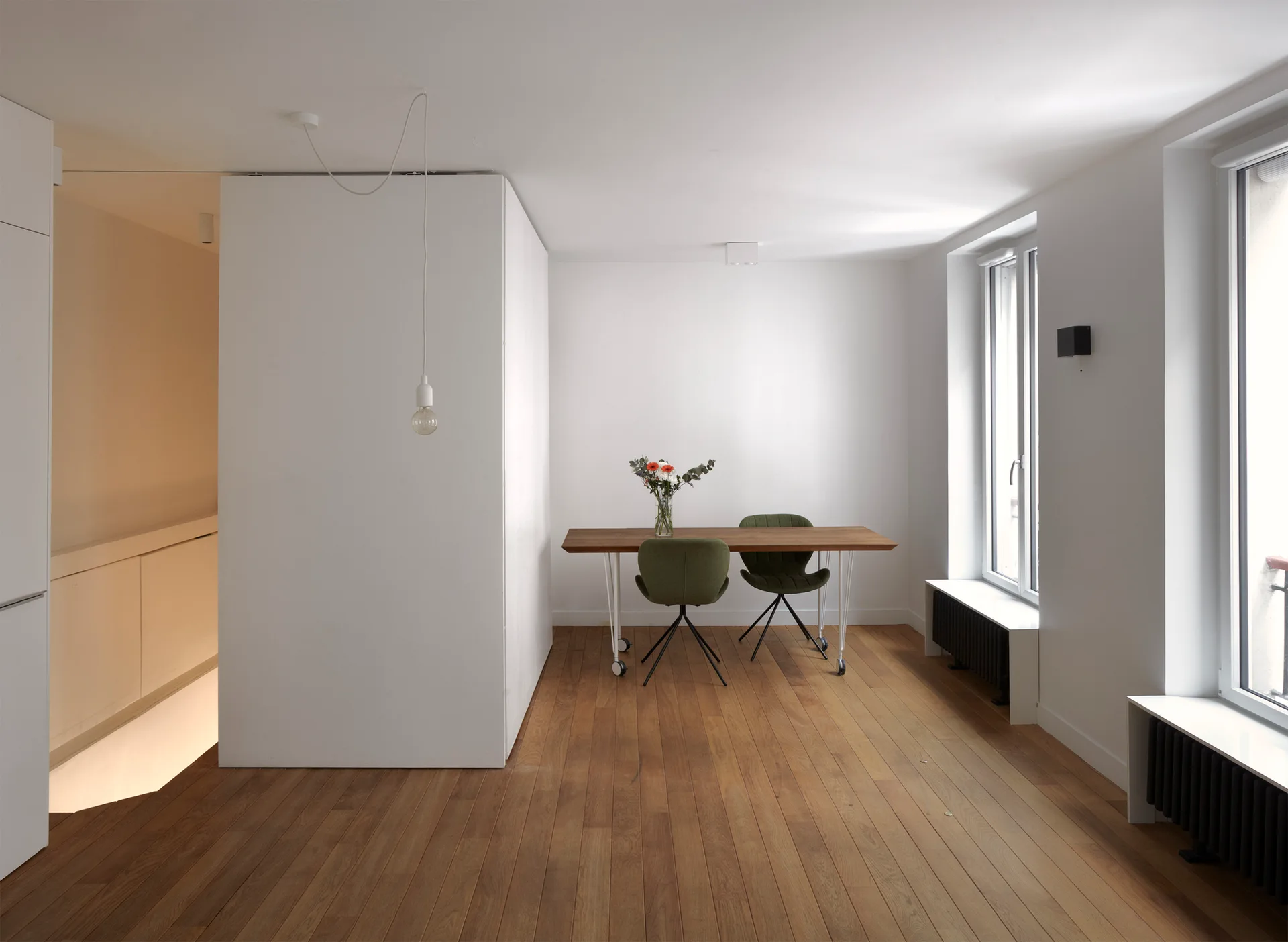 A minimalist dining room features a natural wood table with green upholstered chairs, a white partition wall, and hardwood floors.
