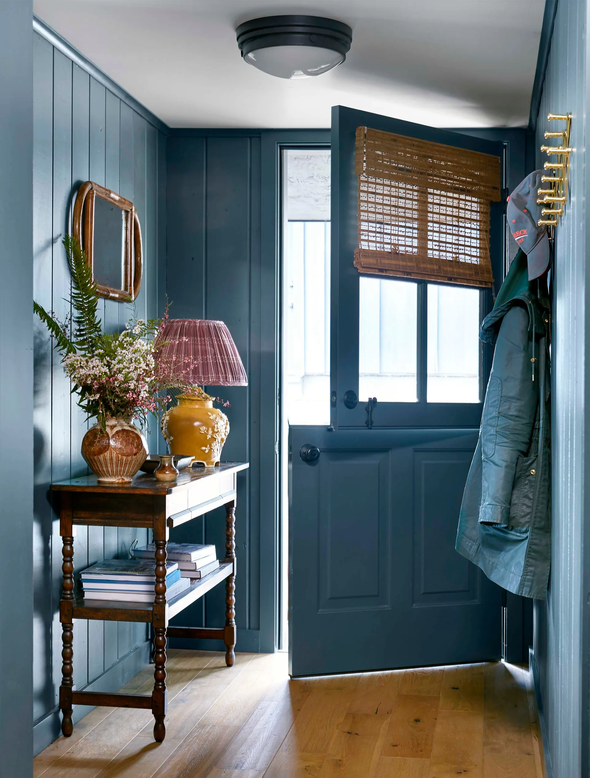 A residential entryway features dark teal vertical wall paneling, a matching open Dutch door, and a classic wood console table decorated with a lamp.