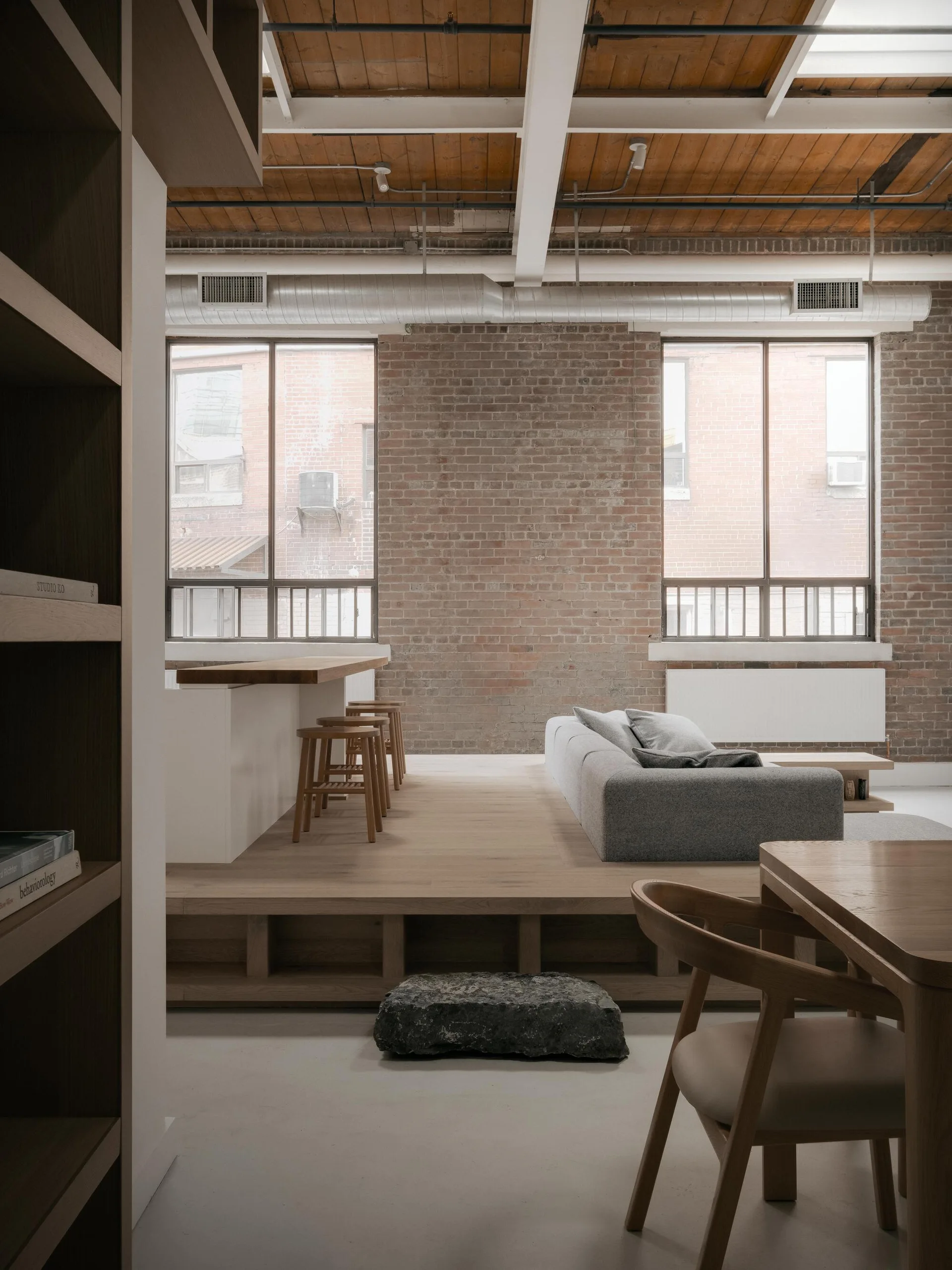 A loft living room features a grey sofa on a raised wood platform, exposed red brick walls, and a timber ceiling with white beams.