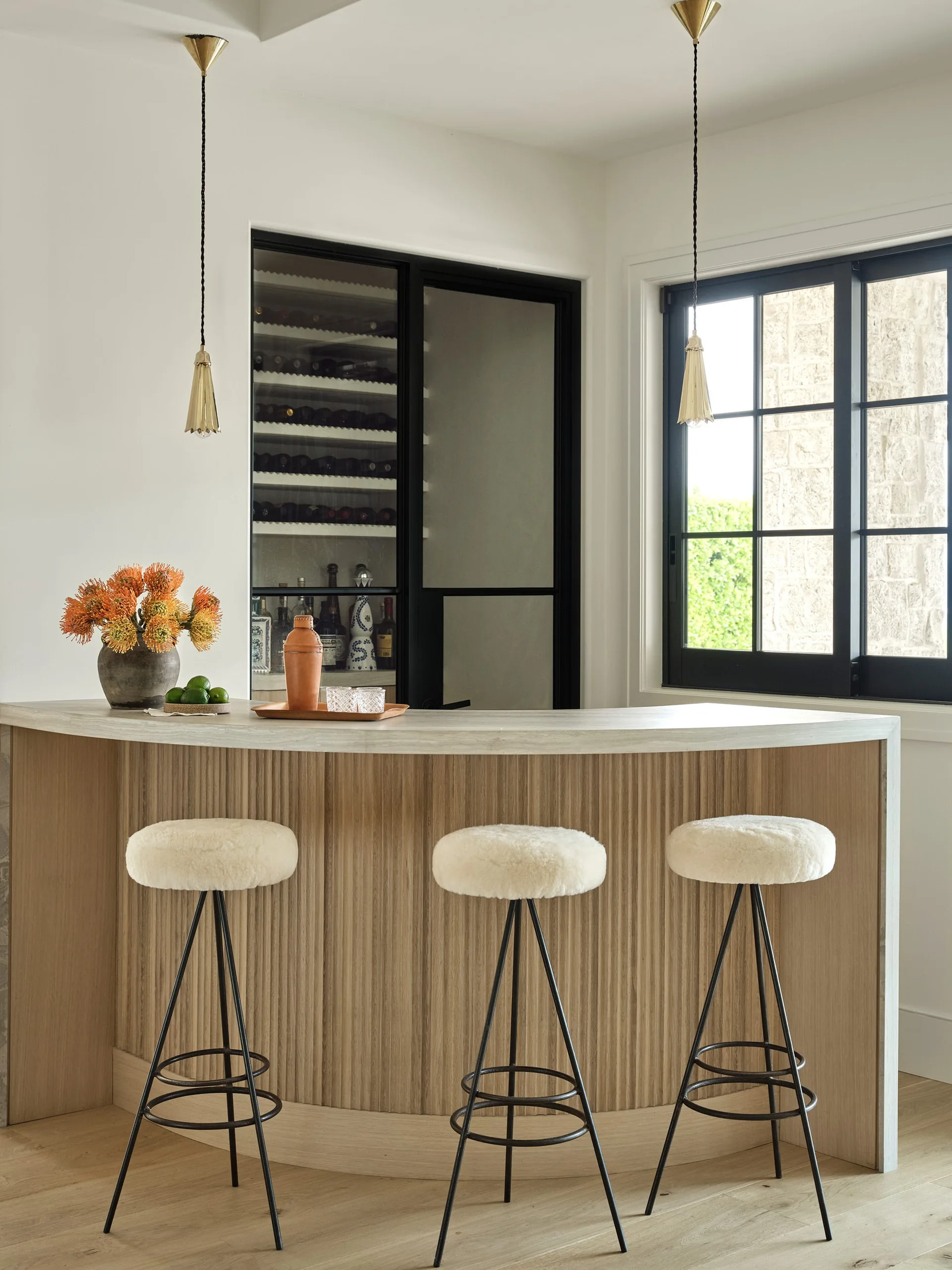 A curved light wood bar counter with vertical ribbing features three fluffy white stools, brass pendant lights, and a black-framed wine cabinet.