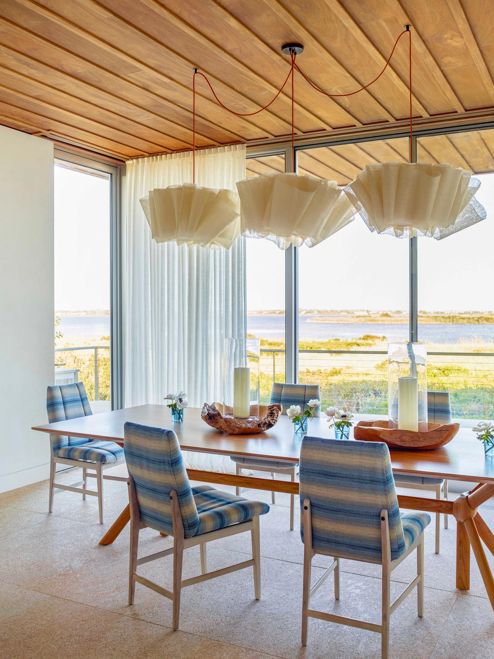 A bright coastal dining room with a long wood table, striped blue chairs, ruffled pendant lights, and floor-to-ceiling windows showing a shoreline view.