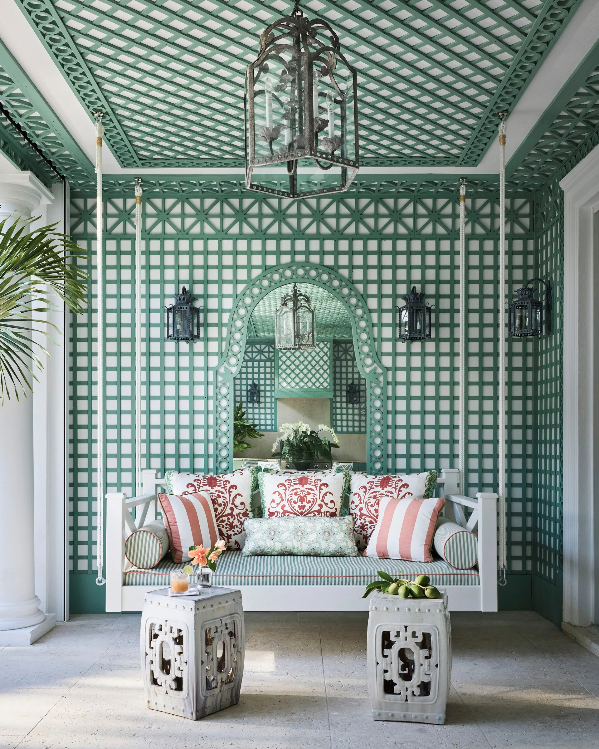 A white hanging swing bench with patterned cushions sits on a porch featuring green wood lattice walls, ceramic garden stools, and a hanging metal lantern.
