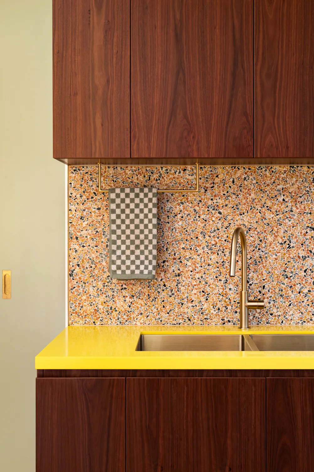 A close-up of a kitchen featuring dark wood cabinets, a bright yellow countertop, a brass faucet, and a multi-colored terrazzo backsplash.