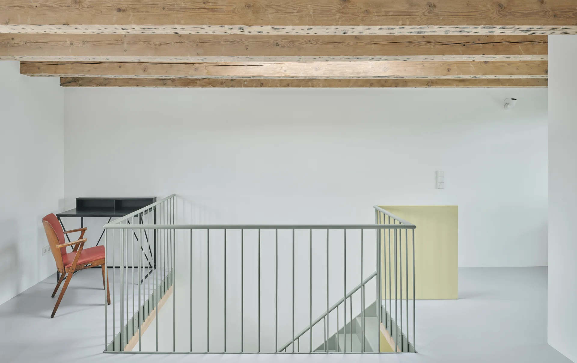 A minimalist home landing featuring exposed raw wood ceiling beams, white walls, and a pale green metal banister surrounding a staircase.