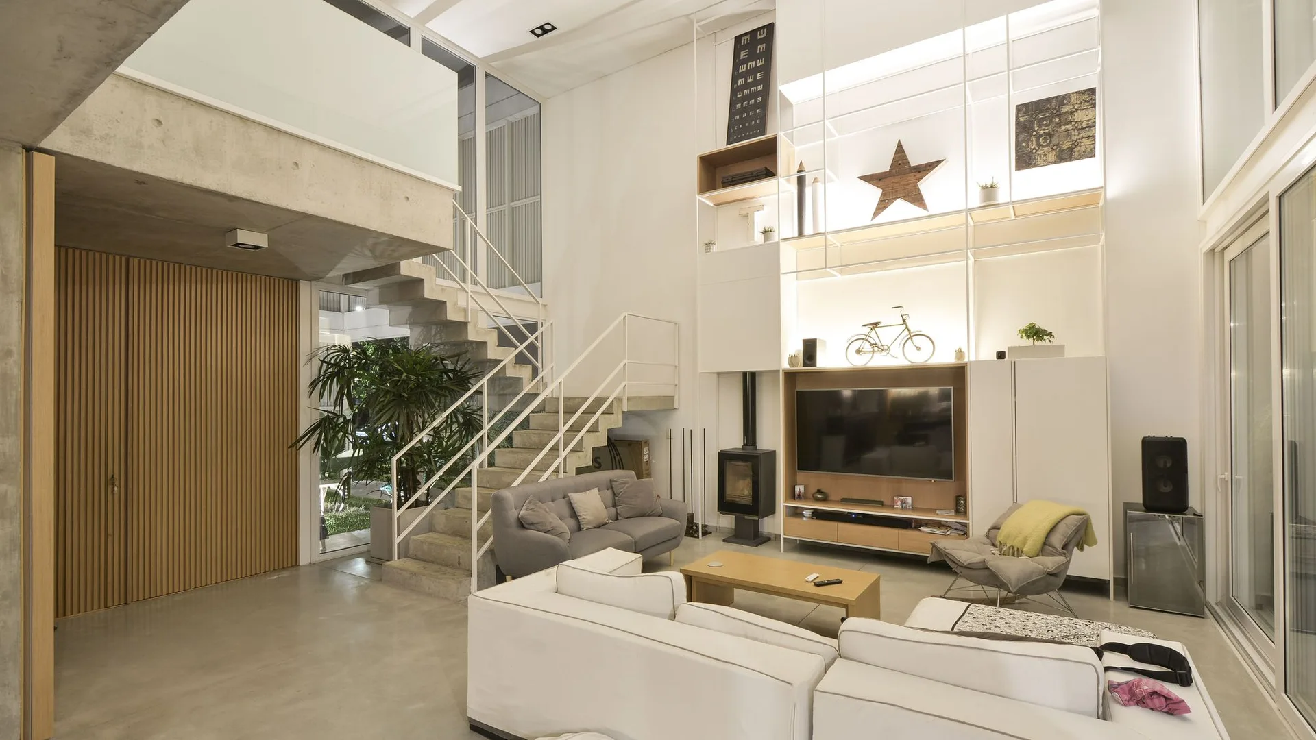 A modern double-height living room featuring grey and white sofas, concrete stairs with a white metal railing, and a tall backlit shelving unit.