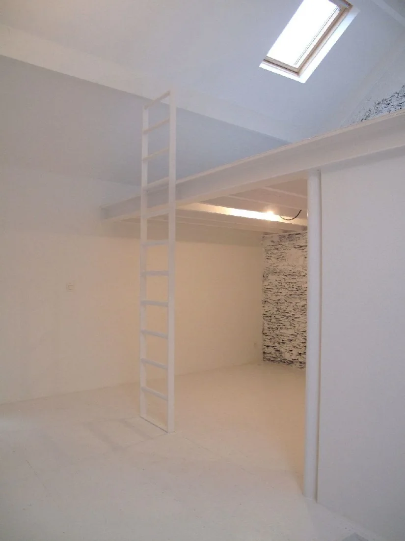 A minimalist white loft room features a vertical metal ladder, a slanted ceiling with a skylight, and a textured white stone accent wall.