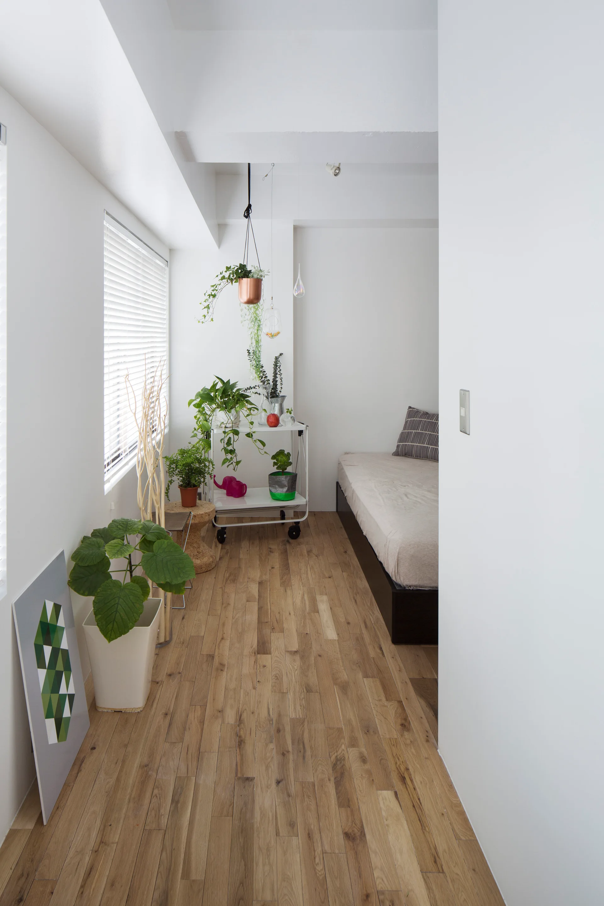 A narrow white bedroom featuring wood plank floors, a low bed, and a rolling white cart filled with green houseplants next to a window.