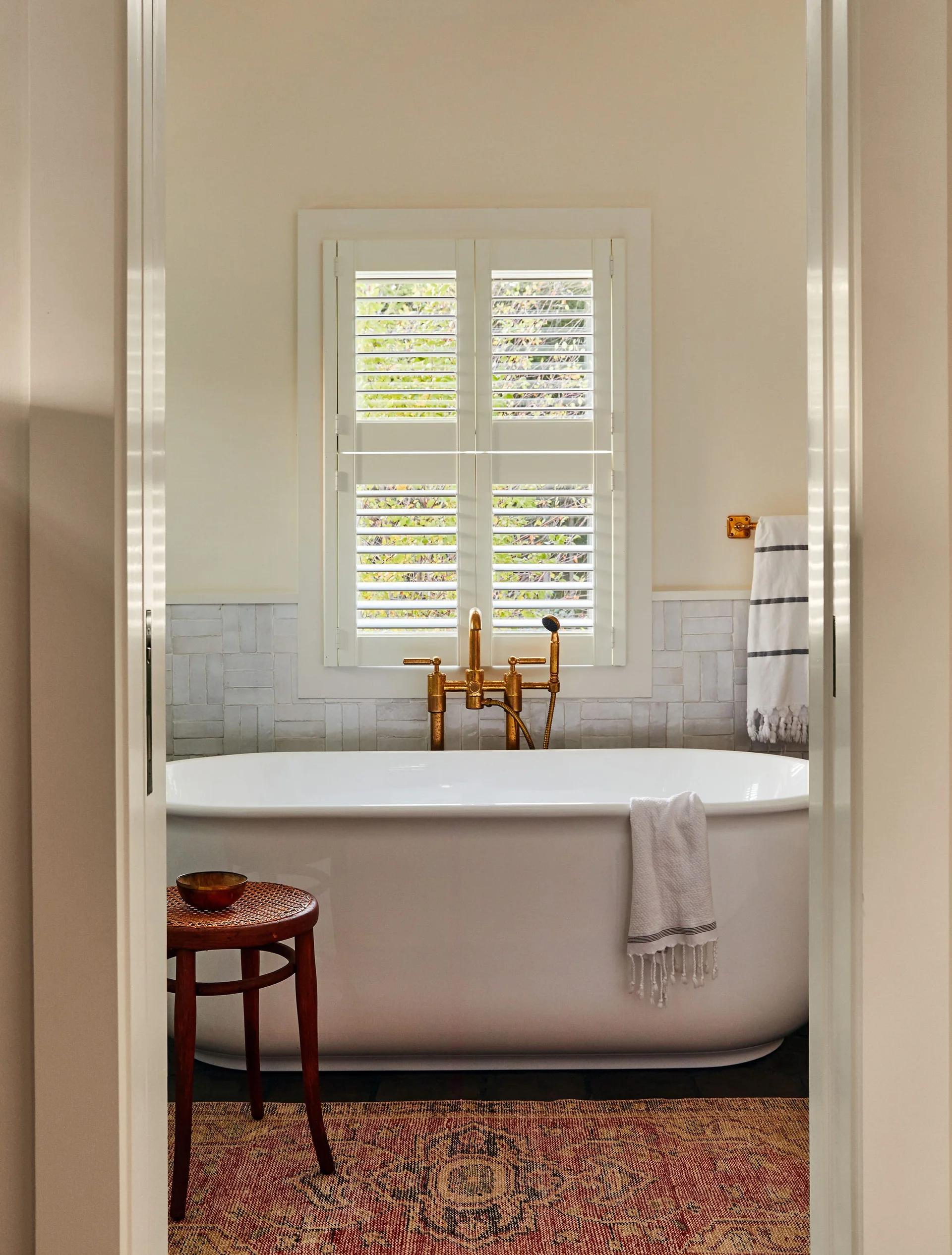 A bathroom view through a doorway featuring a freestanding white soaking tub with brass fixtures, a tiled wall, and a shuttered window.