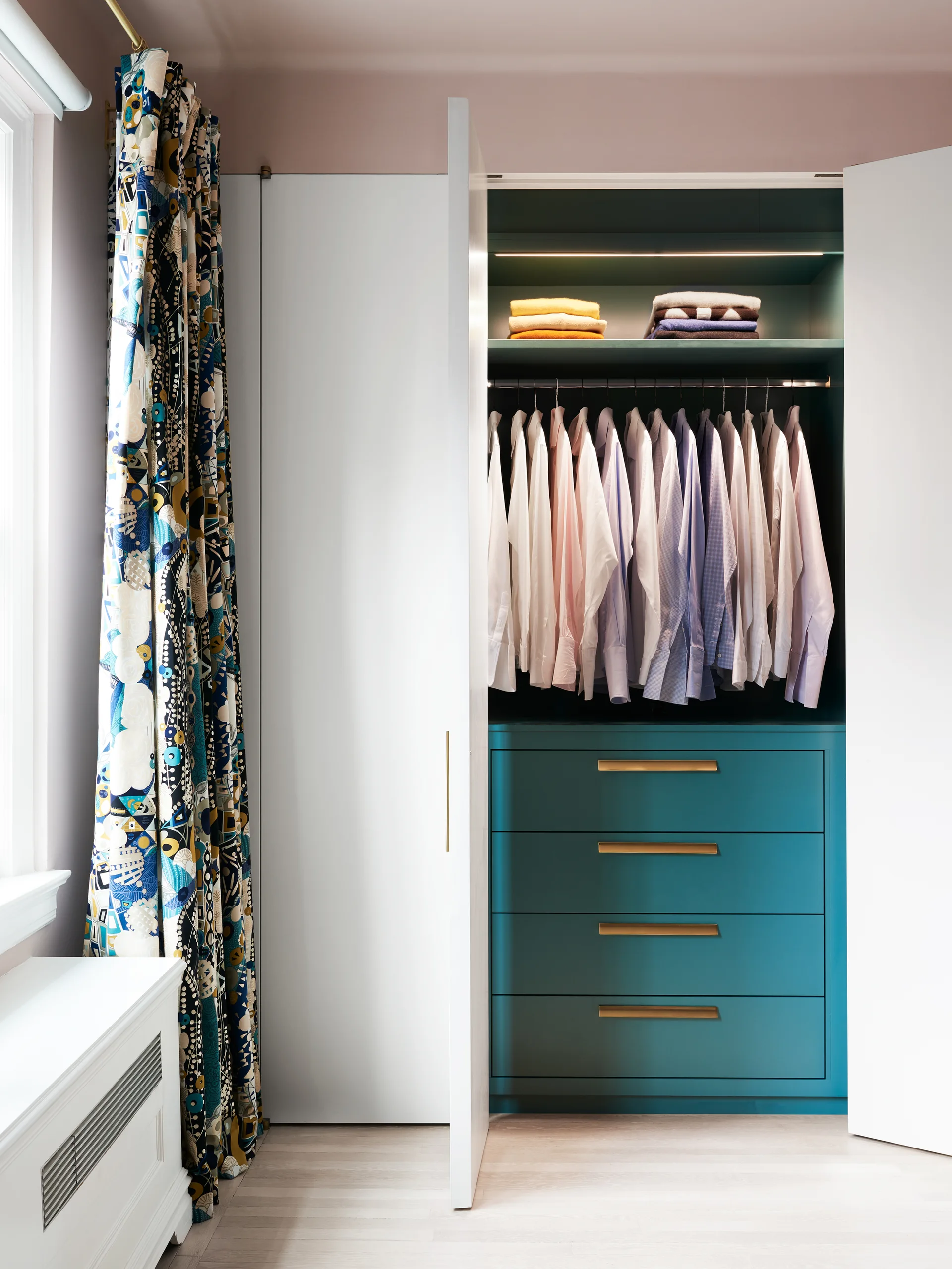 An open white built-in wardrobe features a vibrant teal interior with integrated LED lighting, gold-handled drawers, and hanging shirts.