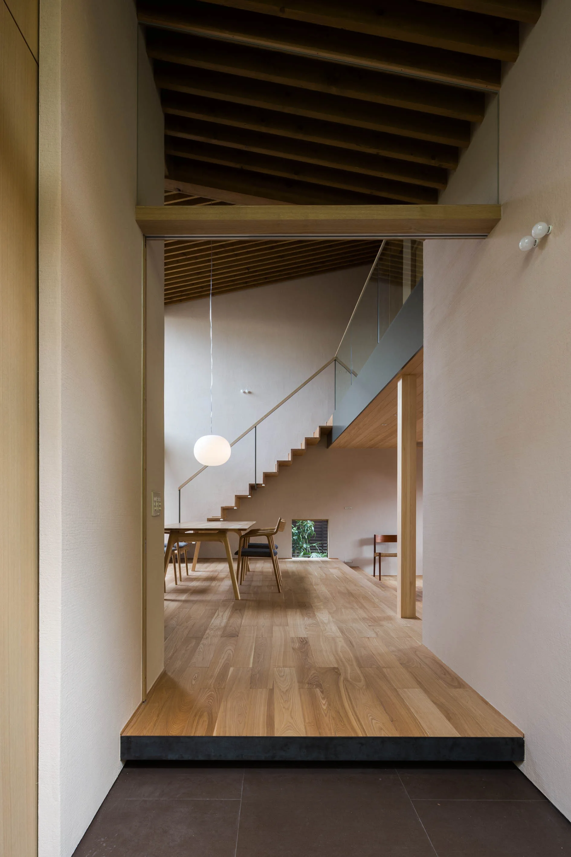 A minimalist wooden dining area viewed from an entryway, featuring a slatted wood ceiling, a floating staircase, and a spherical white pendant light.