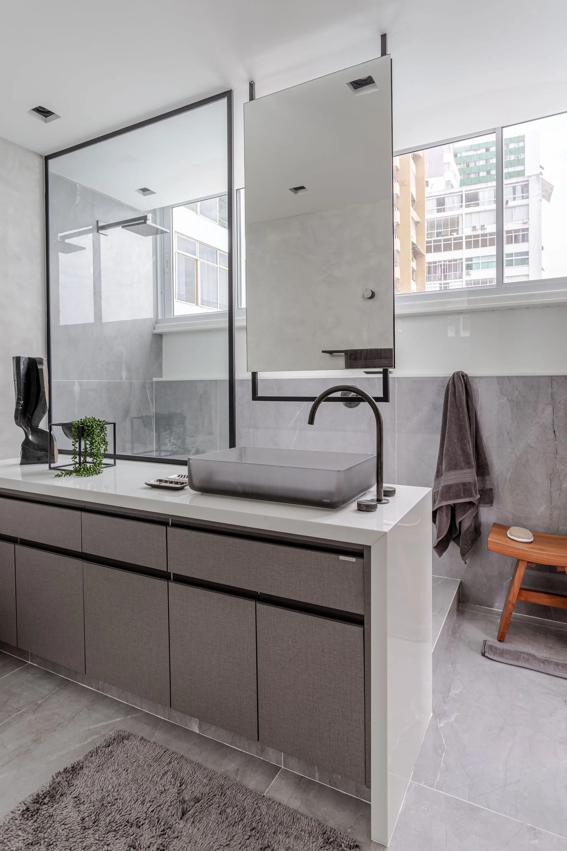 A modern bathroom features a gray textured vanity with a translucent vessel sink, a ceiling-hung mirror, and a glass-enclosed shower.