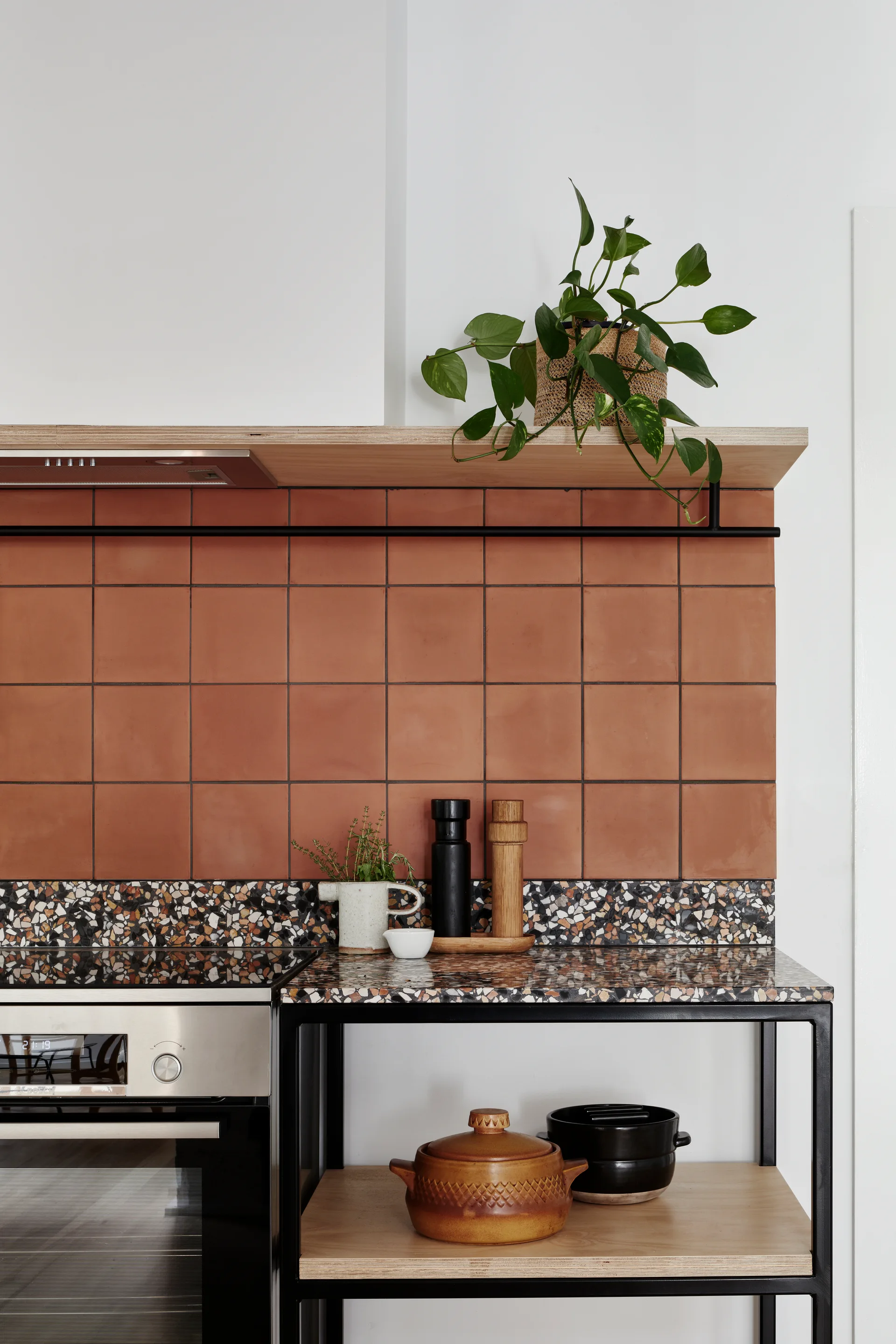 A kitchen detail showcasing a terracotta tile backsplash, a multi-colored terrazzo countertop, and open wood shelving with a black metal frame.