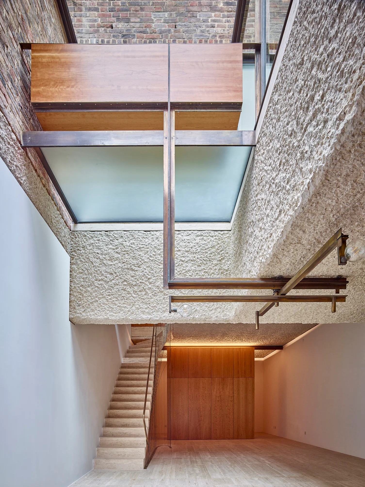 An interior view of a modern stone staircase beneath a glass skylight with heavily textured white walls and warm wood paneling.