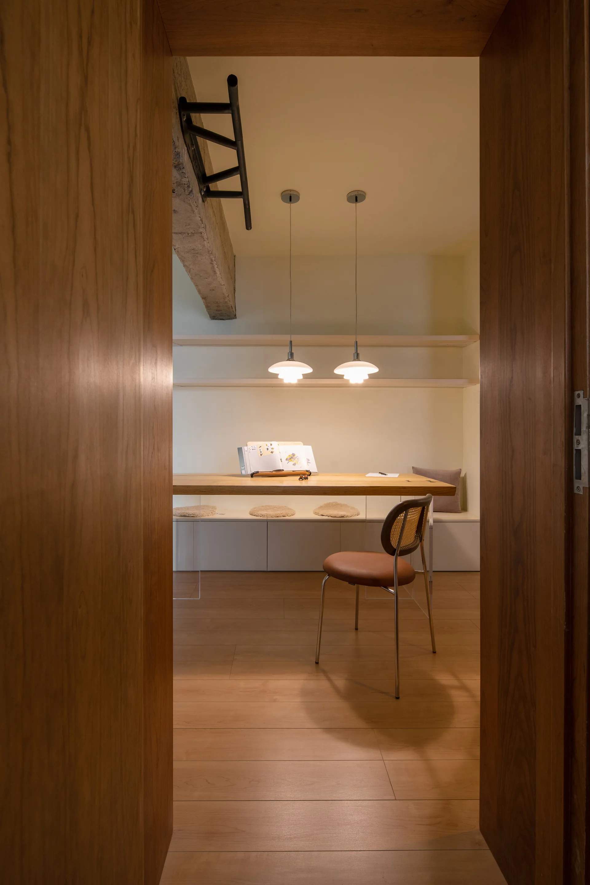 A minimalist study viewed through a wooden doorway features a wood-top desk with clear acrylic legs and a cane-back chair.
