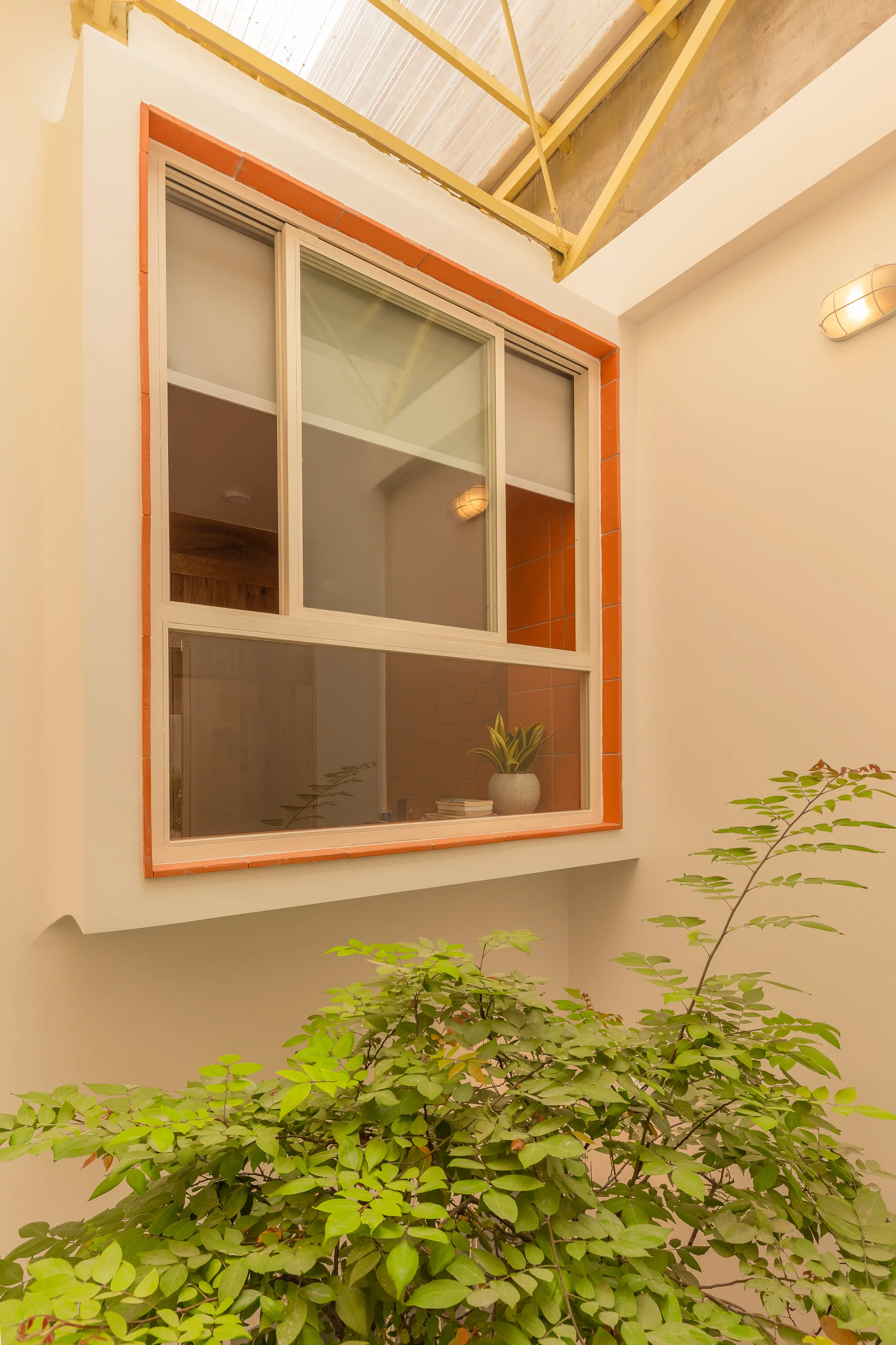 A sliding window framed in bright orange tile sits in a cream-walled courtyard beneath a skylight, with a leafy green plant in the foreground.