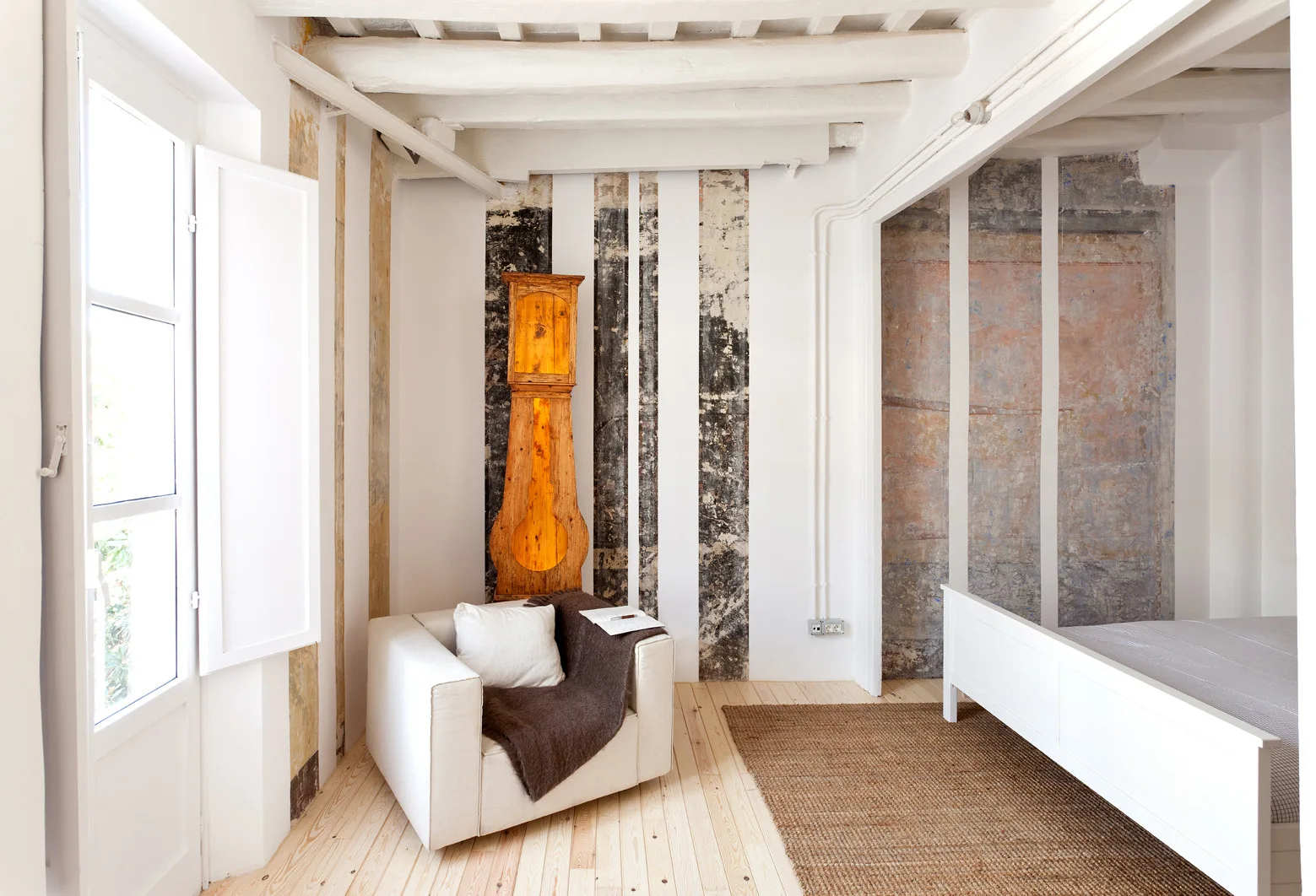 A bright bedroom with exposed white ceiling beams, a wooden grandfather clock, a white armchair, and vertical structural detailing on the walls.
