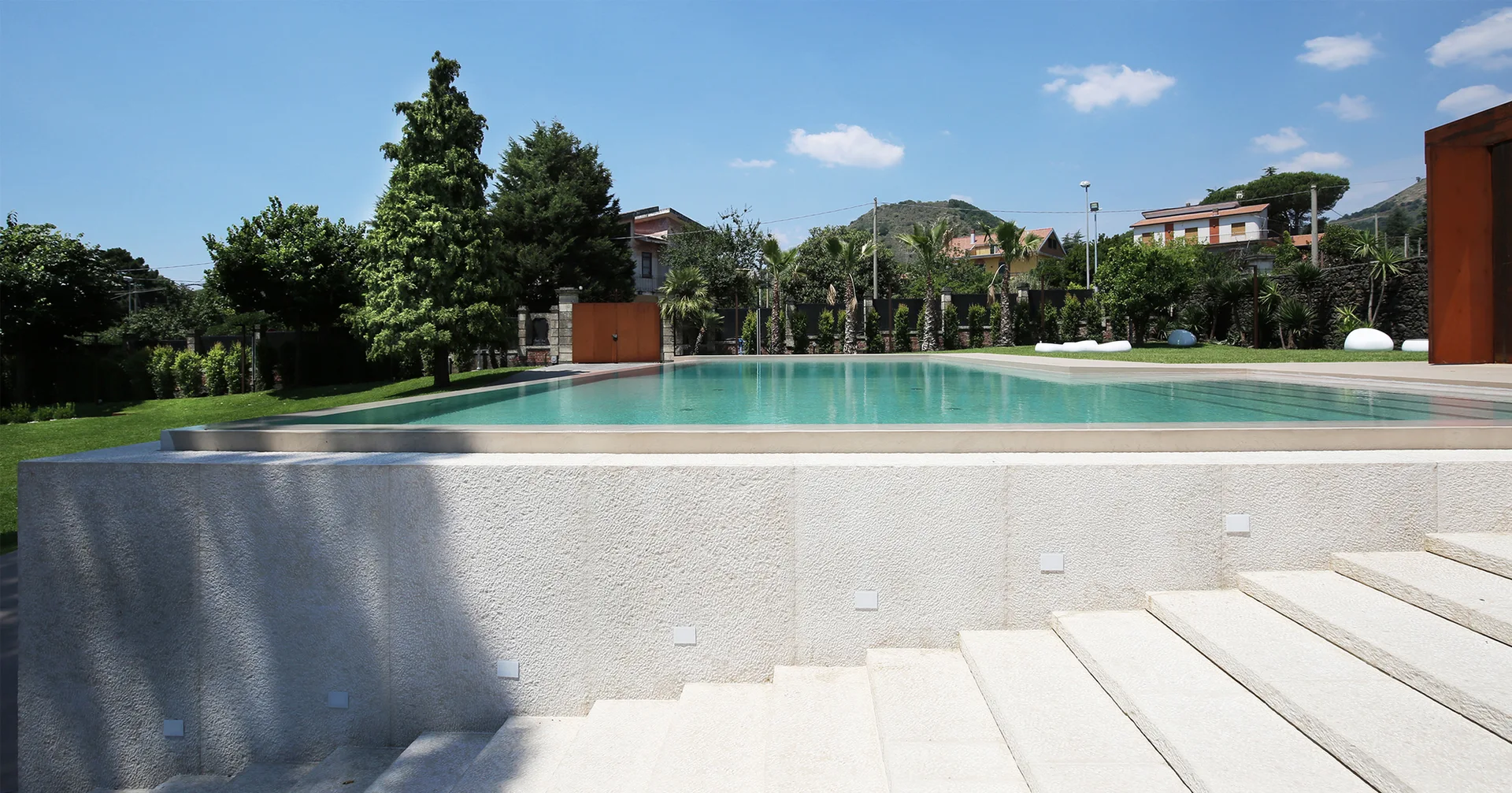 Wide textured stone steps with integrated square lights lead up to a rectangular swimming pool set in a grassy backyard under a blue sky.