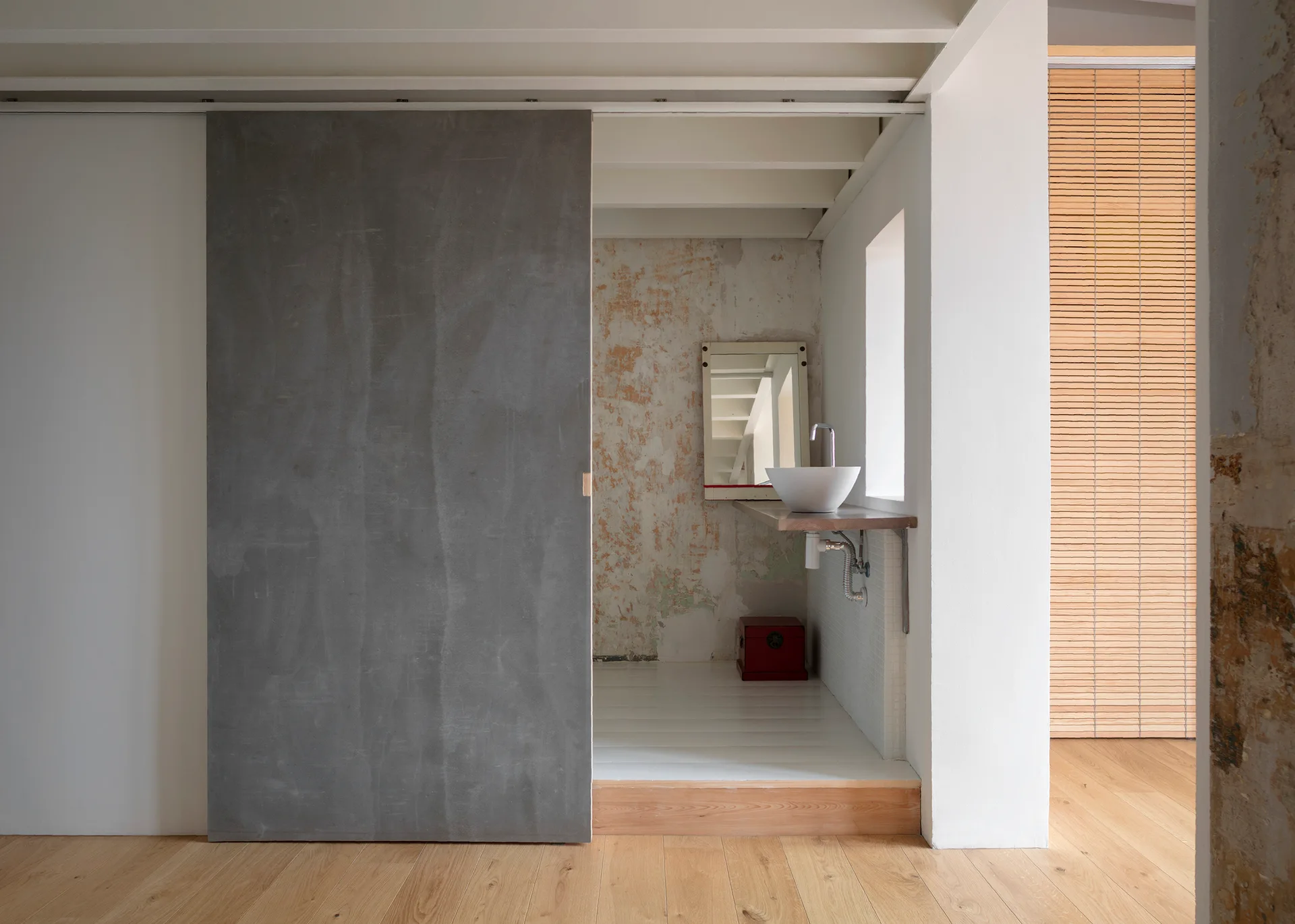A minimalist bathroom alcove featuring a grey sliding door, a white vessel sink on a floating wood vanity, and a distressed plaster accent wall.