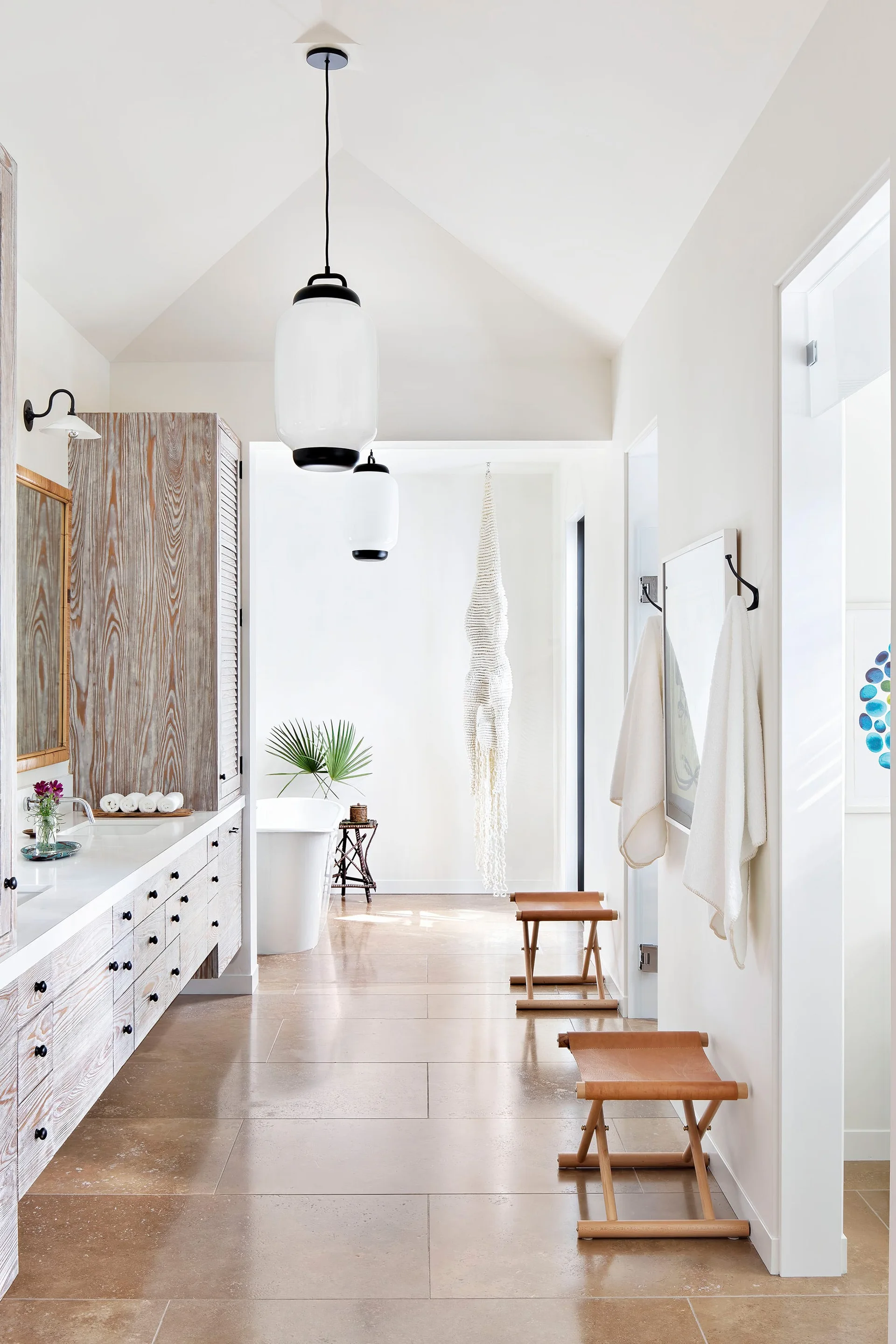 A bright bathroom features a long light-wood double vanity, large beige floor tiles, two white pendant lights, and a freestanding bathtub.