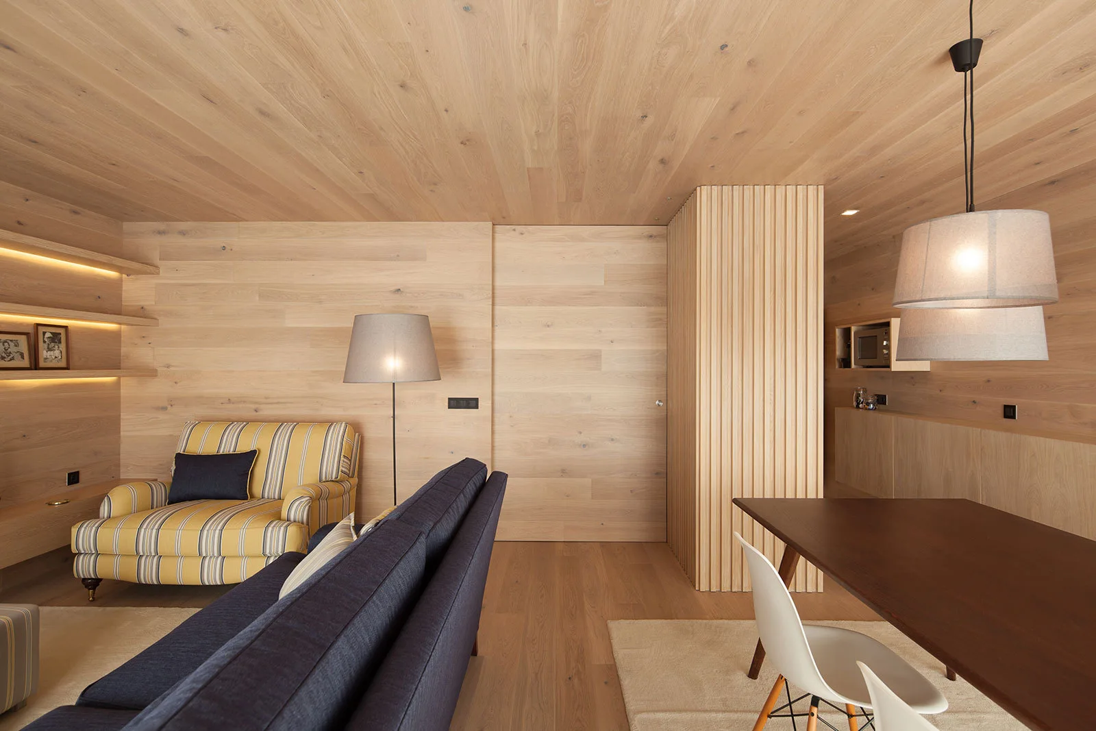 A wood-paneled living room featuring a dark blue sofa, a yellow striped armchair, a slatted wooden pillar, and a dining table.